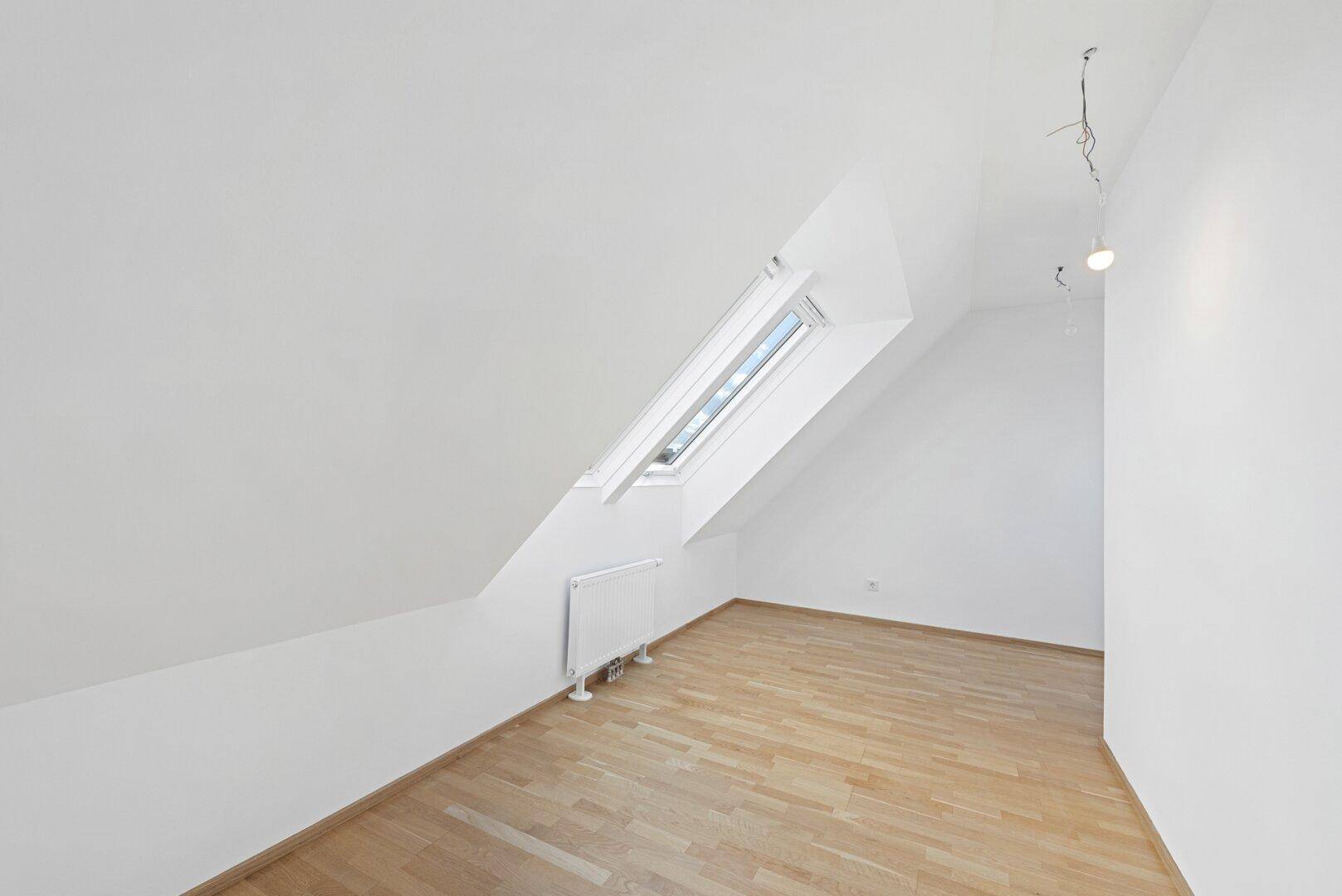 Helles Zimmer mit Holzboden, Dachschräge und einem einzelnen Dachfenster.