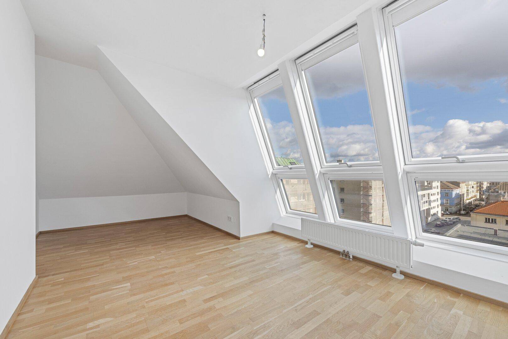 Helles Zimmer mit Holzboden, mehreren Dachfenstern und weitem Stadtblick.