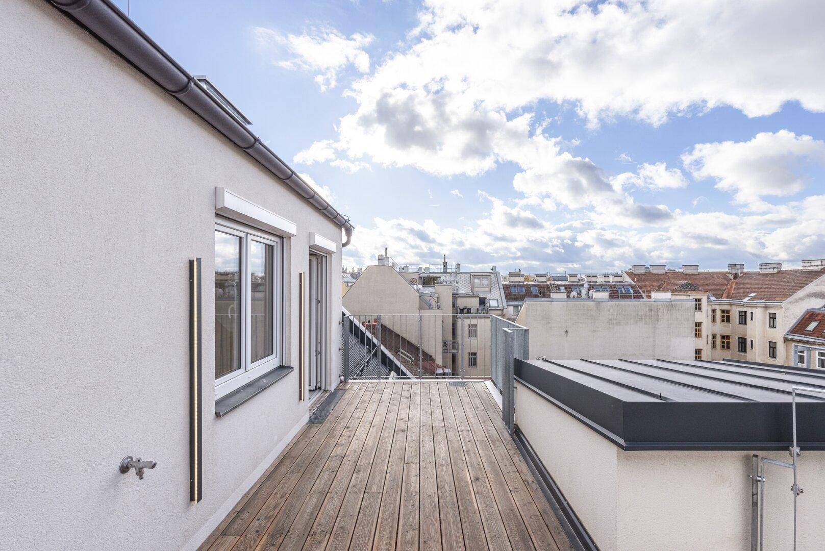 Dachterrasse mit Holzboden, weißer Fassade und Blick auf die umliegenden Gebäude.