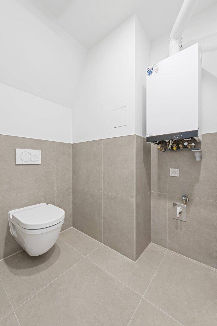 Kleiner, modern gefliester WC-Raum mit wandhängender Toilette und Warmwasserbereiter.