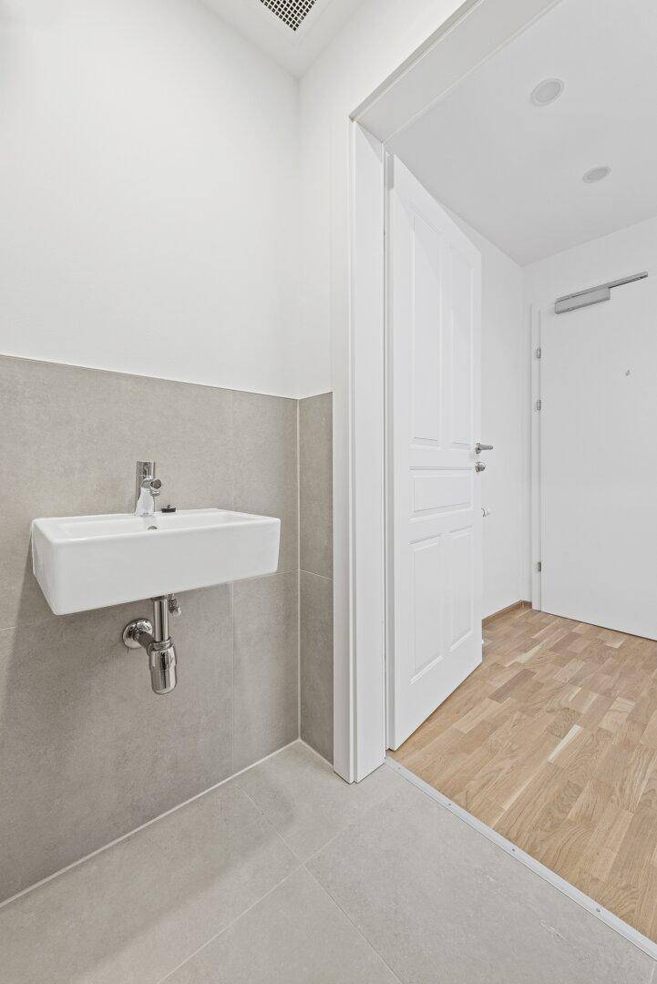 Modernes WC mit wandmontiertem Waschbecken und hellen Fliesen, angrenzend an einen Holzfußboden.