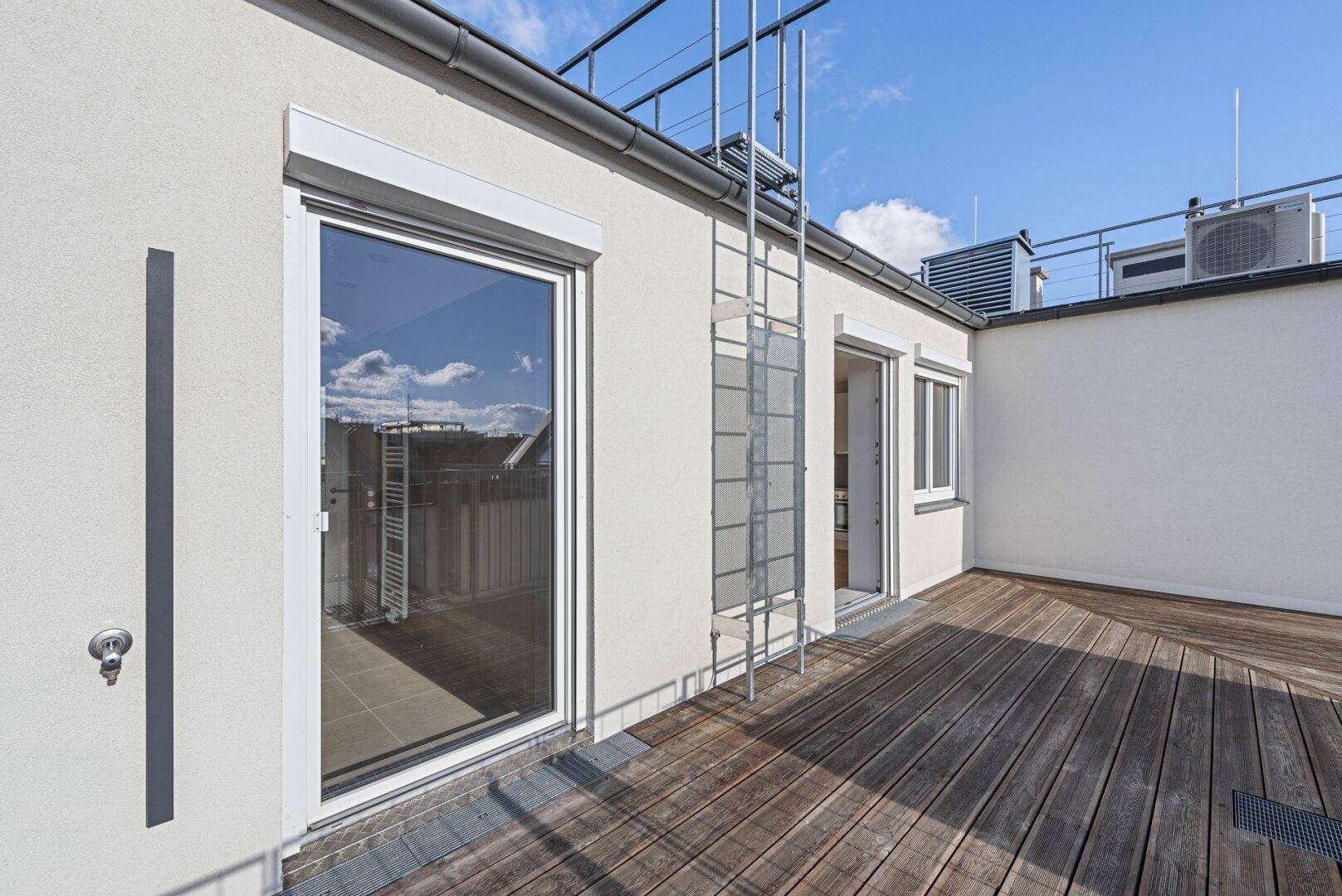 Helle Dachterrasse mit Holzboden, großen Fenstern und Zugang zu den Innenräumen des Gebäudes.
