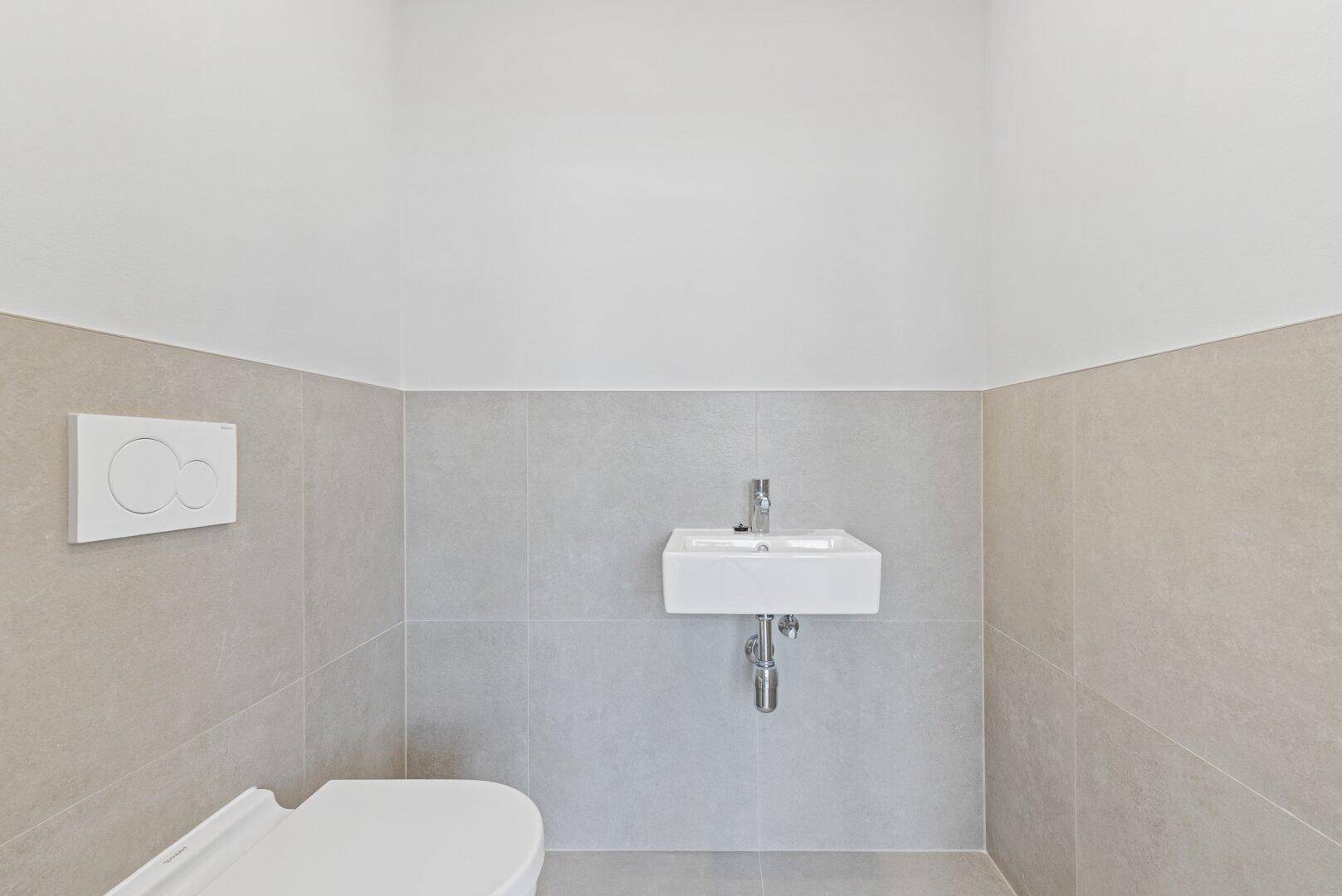 Modernes WC mit wandhängender Toilette und Waschbecken in einem hell gefliesten Raum.