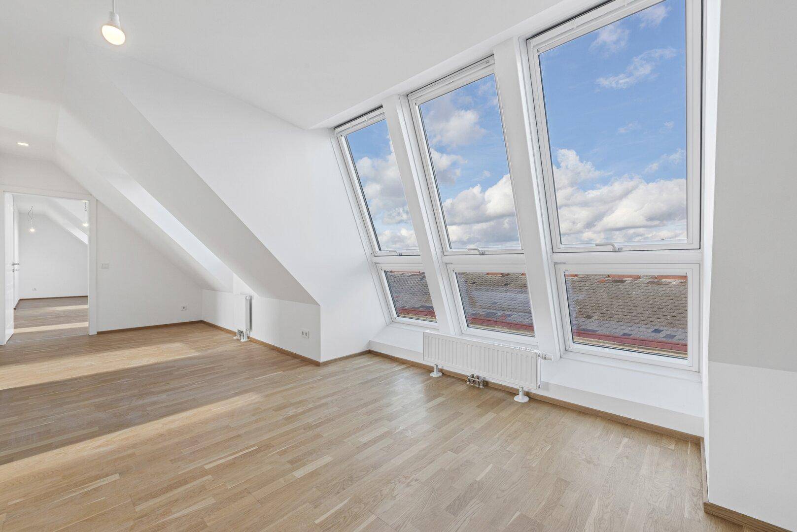 Heller Dachgeschossraum mit schrägen Wänden, Holzboden und großen Dachfenstern mit Stadtblick.