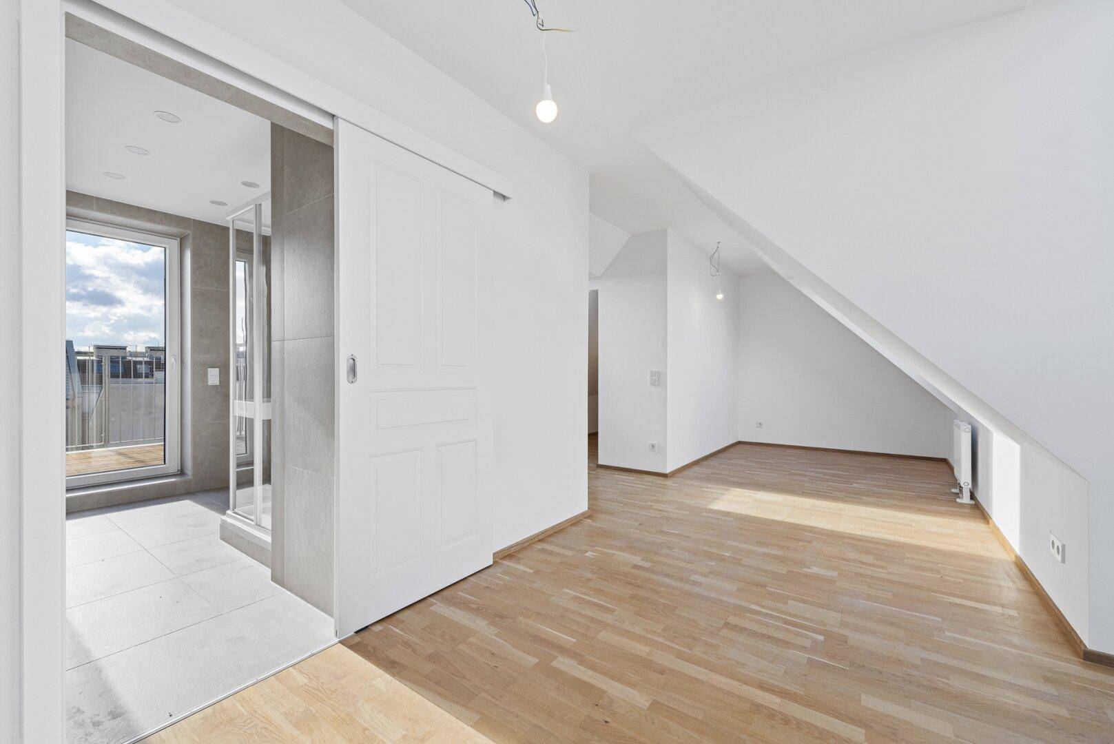 Heller Raum mit Holzboden und Schiebetür, die zu einem modernen Badezimmer mit Dusche führt.