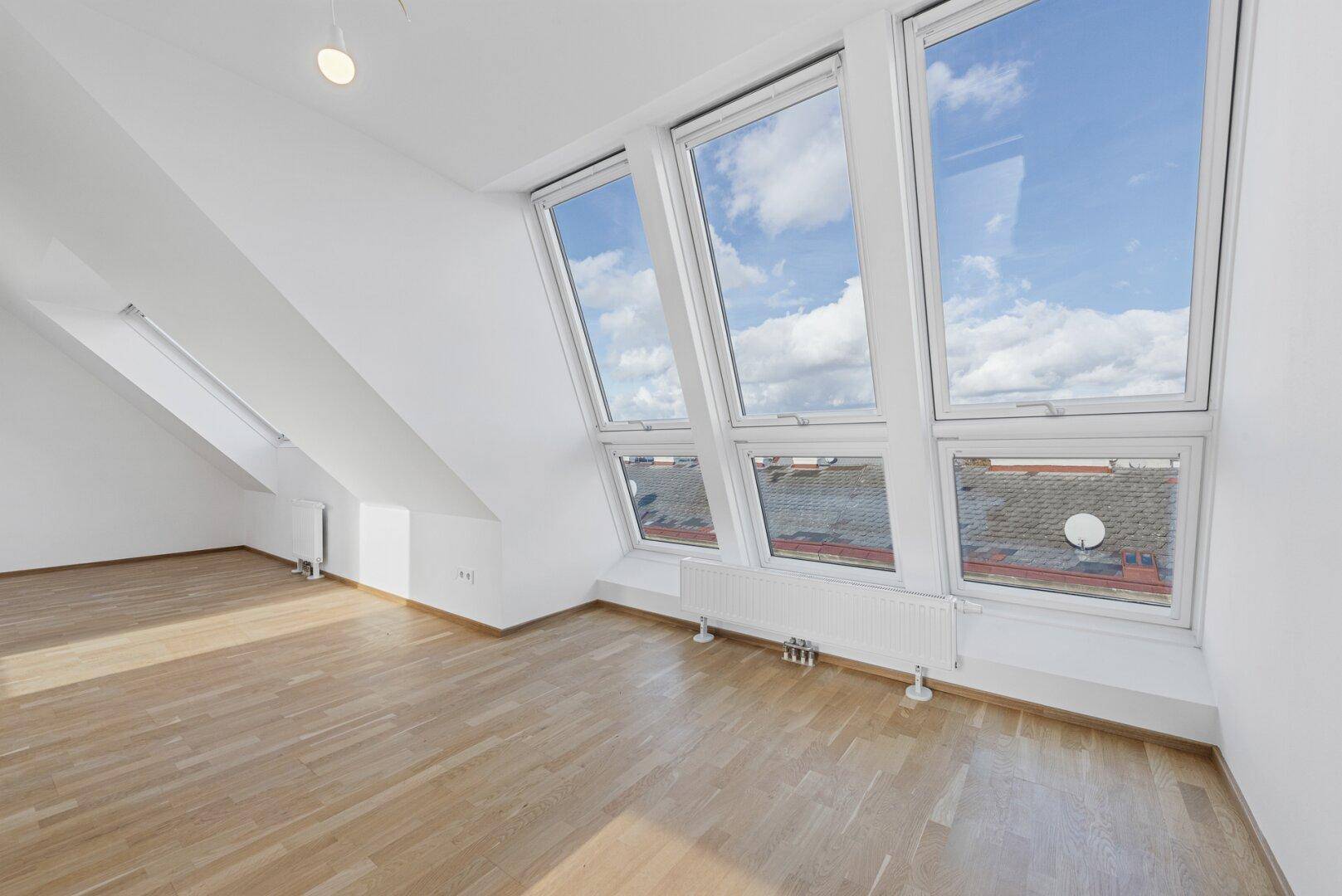 Heller Dachgeschossraum mit schrägen Wänden, Holzboden und großen Dachfenstern mit Stadtblick.