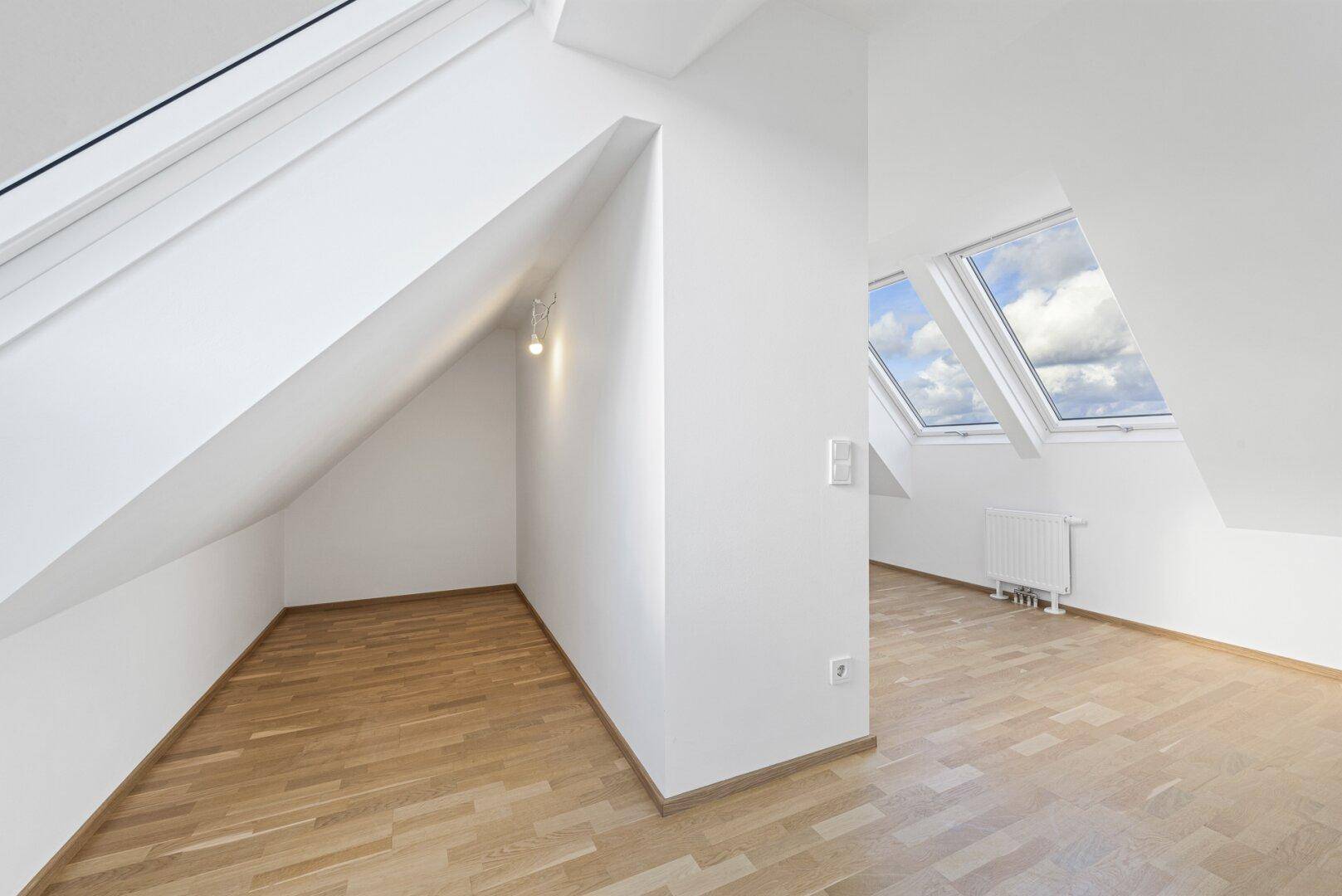 Heller Dachgeschossraum mit Holzboden, schrägen Wänden und zwei Dachfenstern, die Tageslicht spenden.