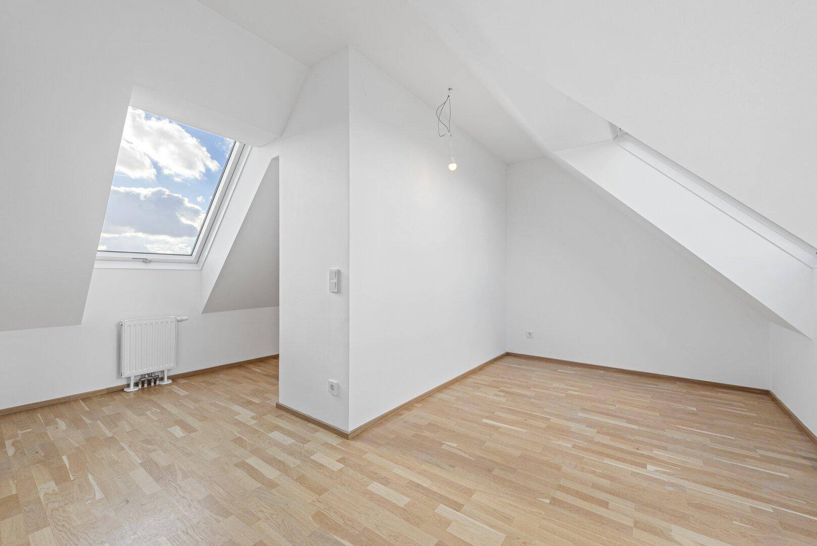 Lichtdurchfluteter Dachgeschossraum mit Holzboden, schrägen Wänden und einem großen Dachfenster.
