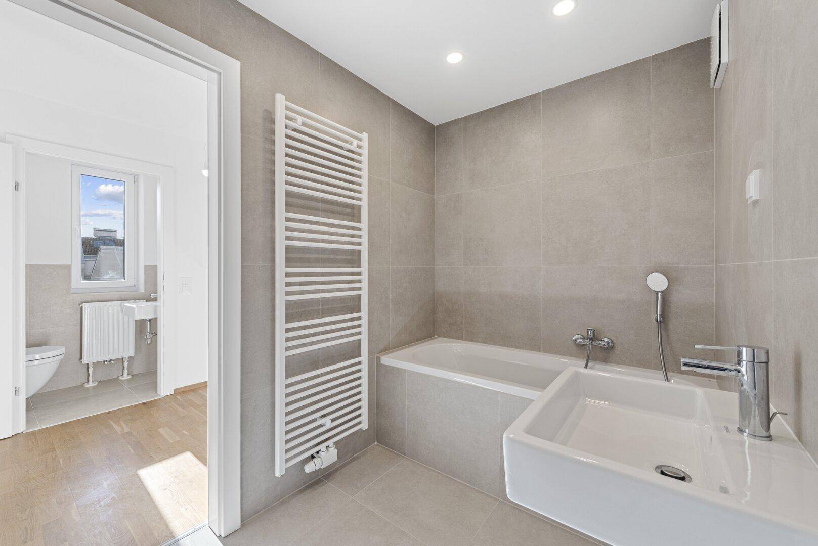 Helles Badezimmer mit Badewanne, Waschbecken und beheiztem Handtuchhalter, mit Blick in ein weiteres Zimmer.