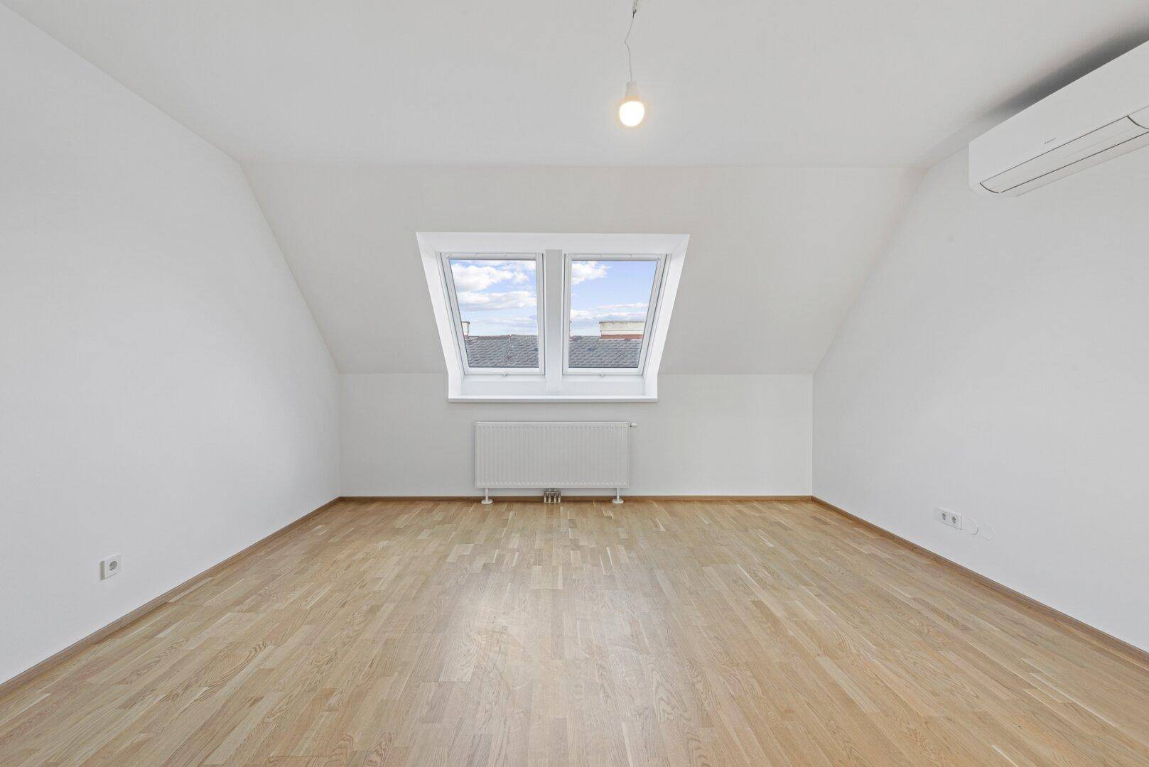 Helles Zimmer mit Parkettboden, schrägen Wänden und zwei Dachfenstern, die viel Licht spenden.