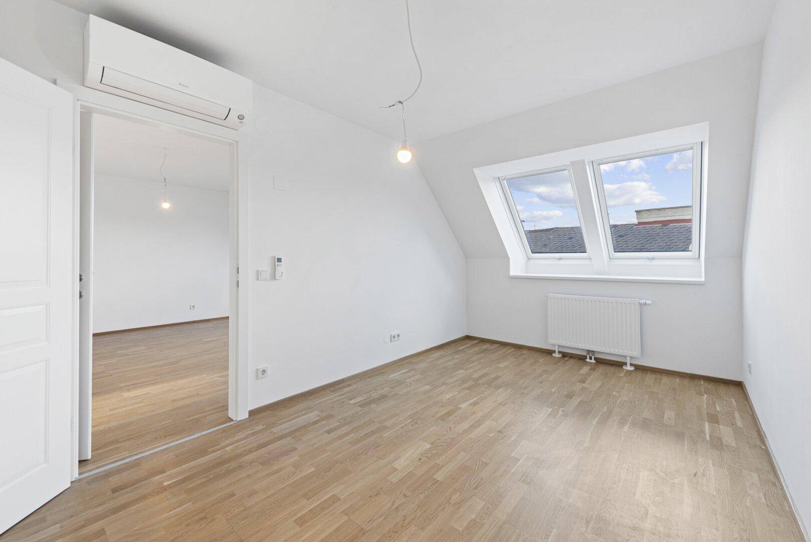 Helles Zimmer mit Parkettboden, Klimaanlage und Dachfenstern, das zu einem weiteren Raum führt.