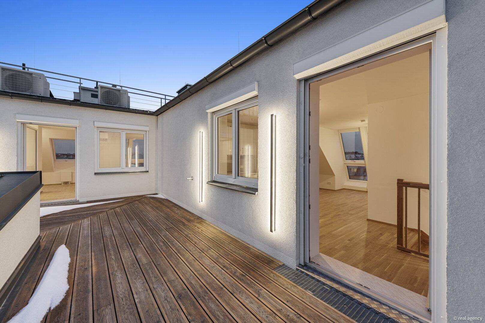 Dachterrasse bei Dämmerung mit Holzboden, beleuchteter Fassade und Stadtblick.