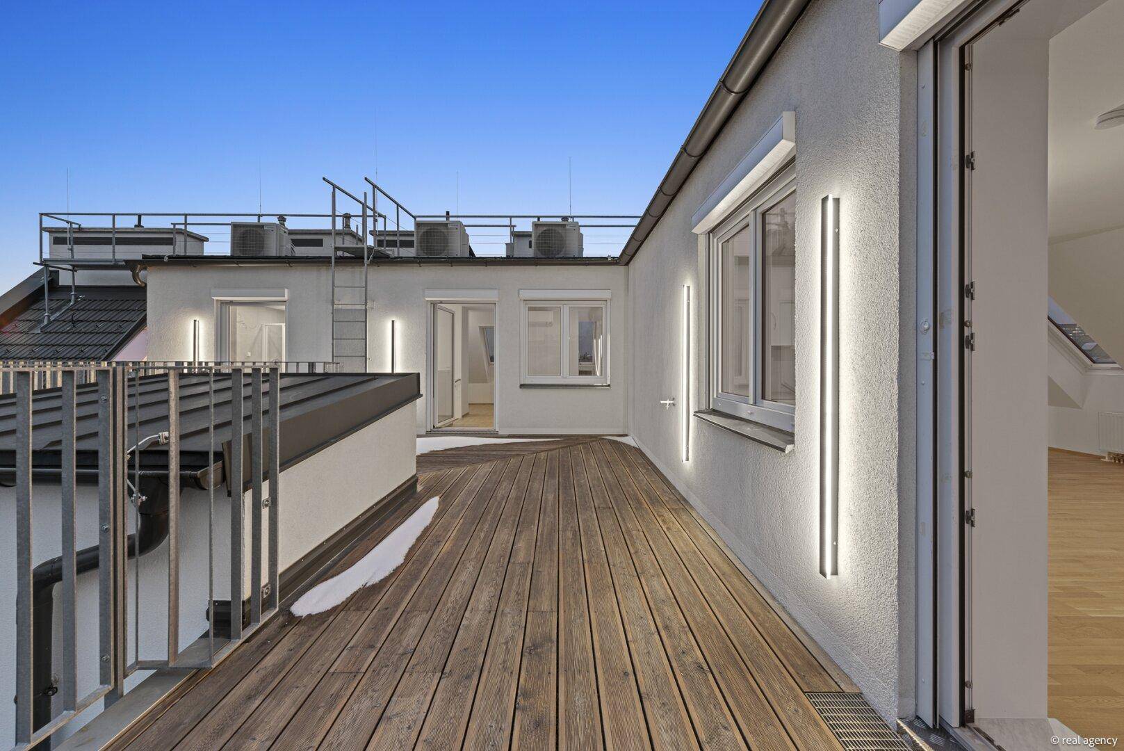 Dachterrasse bei Dämmerung mit Holzboden, modernem Geländer und beleuchteter Fassade.