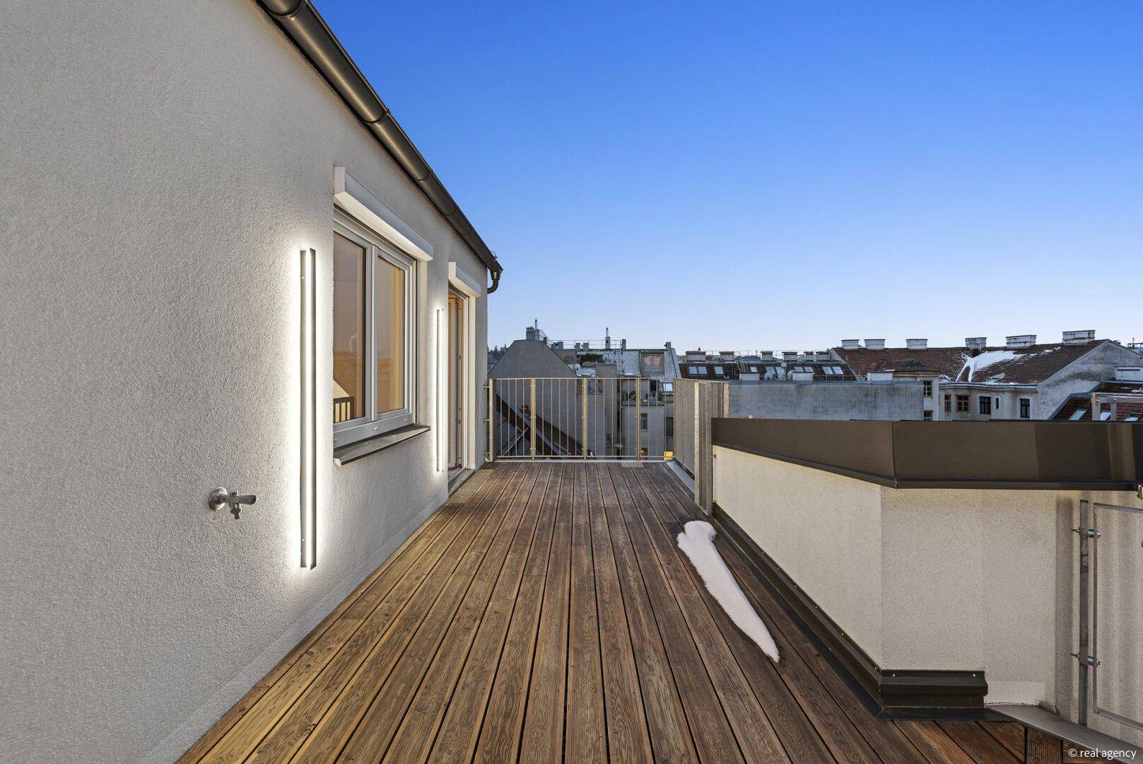 Dachterrasse bei Dämmerung mit Holzboden, beleuchteter Wand und Stadtblick.