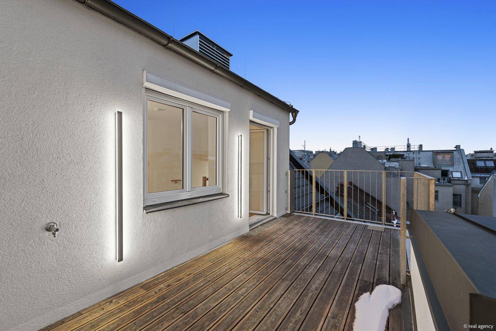 Dachterrasse bei Dämmerung mit Holzboden, beleuchteter Fassade und Zugang zum Innenraum.