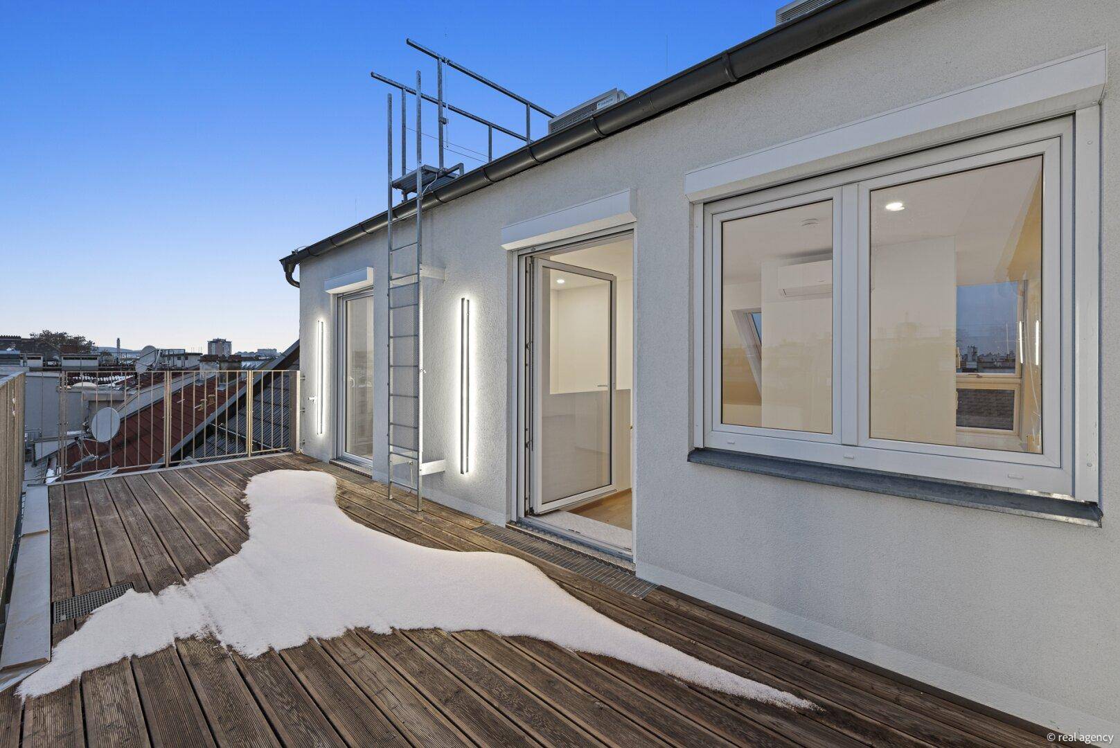 Abendliche Dachterrasse mit Holzboden, moderner Beleuchtung und Blick auf die Stadt bei Dämmerung.