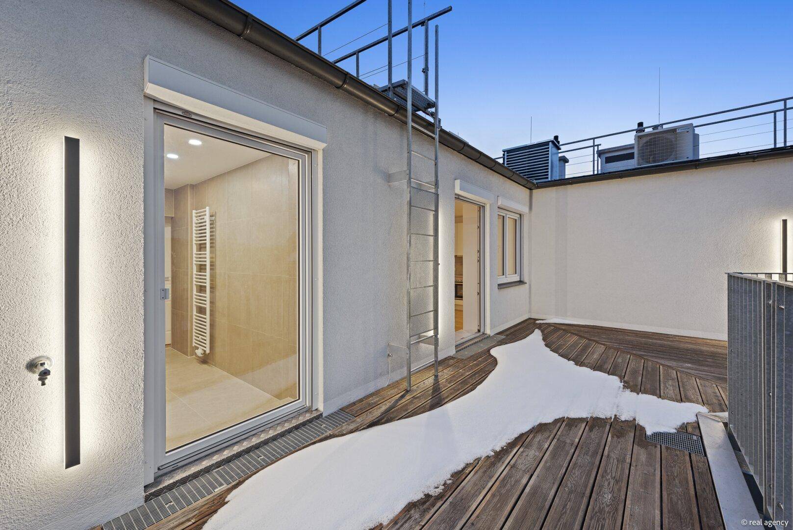 Abendliche Dachterrasse mit Holzboden und Beleuchtung, mit Blick in ein helles Badezimmer.