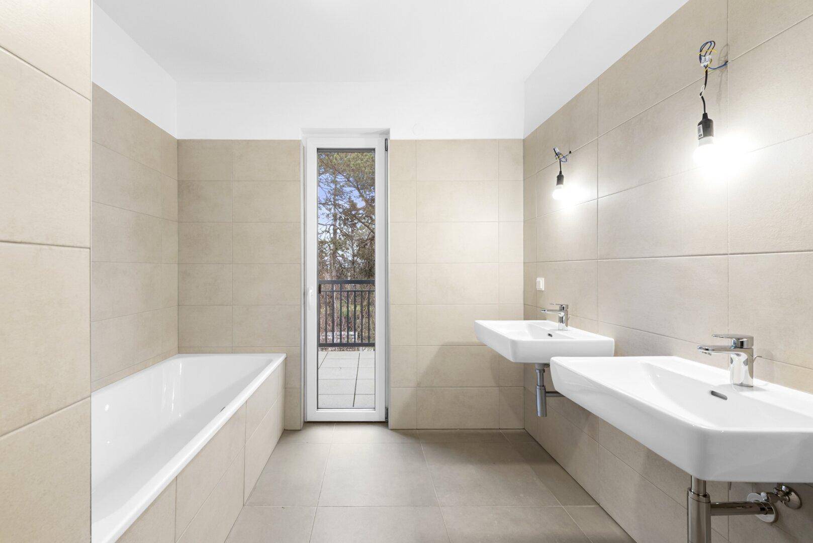 Helles Badezimmer mit Badewanne, Doppelwaschbecken und Balkonzugang.