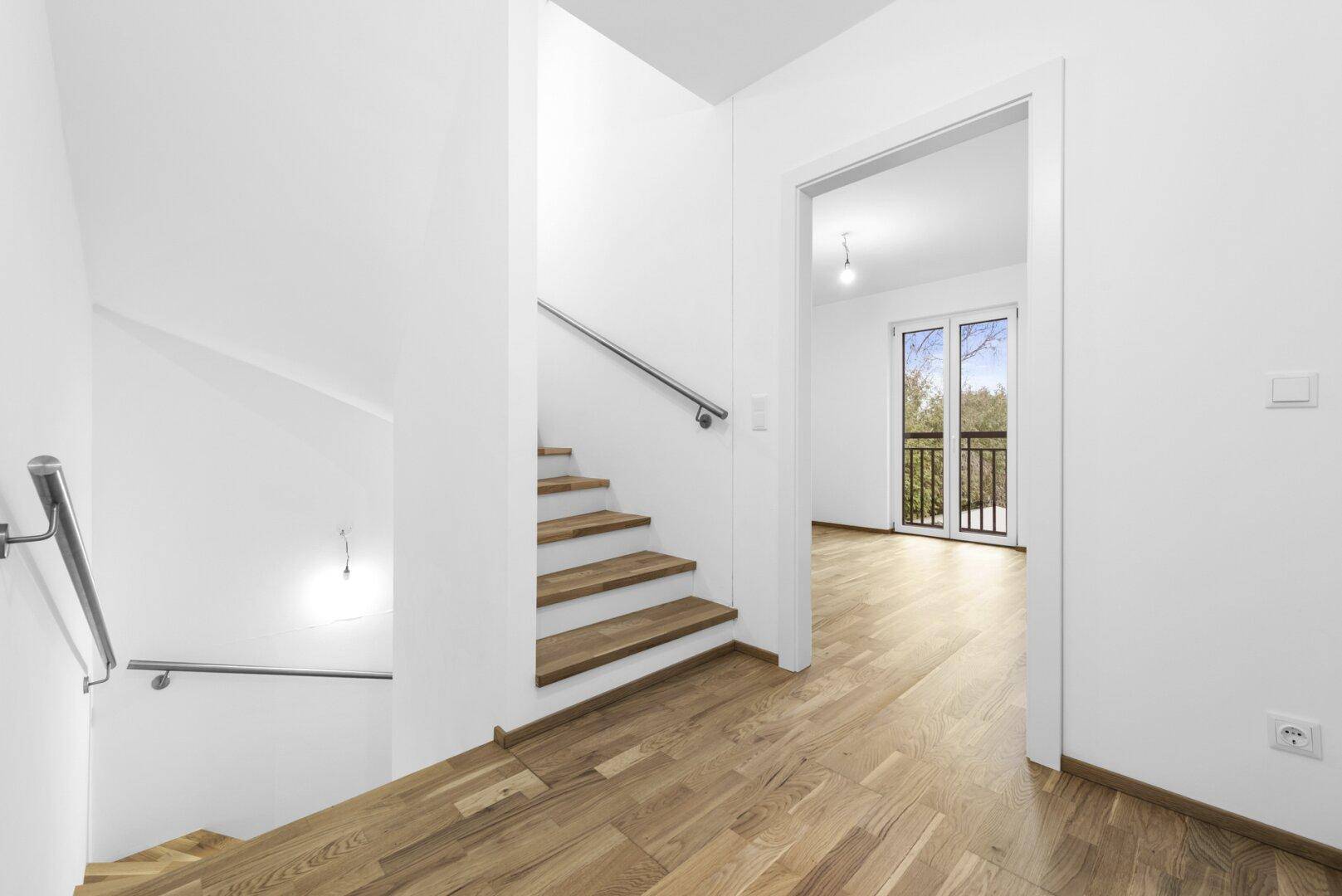 Heller Flur mit Holztreppe und Zugang zu einem Zimmer mit Balkon.