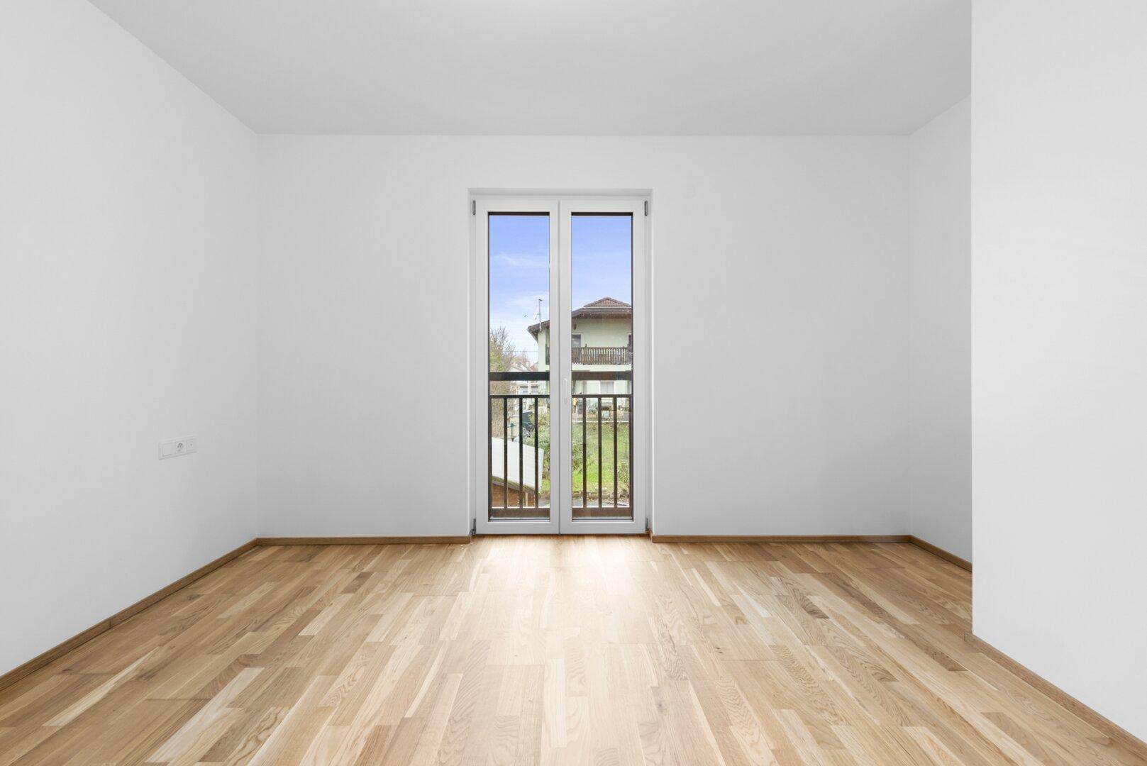 Leeres Zimmer mit Holzboden und französischem Balkon, bietet viel Tageslicht.