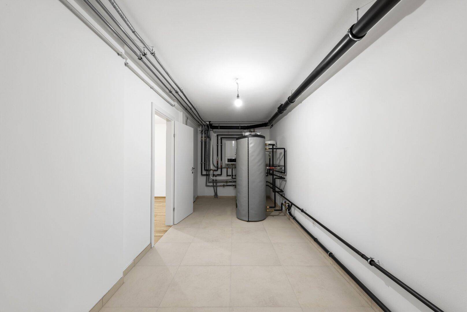Langer Technikraum mit Warmwasserspeicher und sichtbaren Rohrleitungen.
