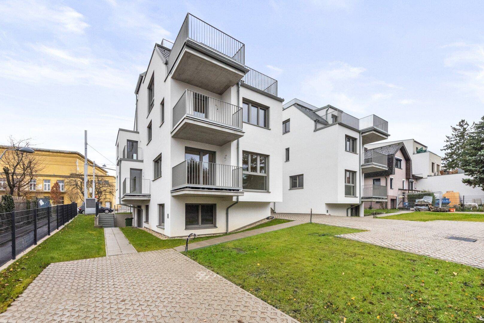 Modernes weißes Mehrfamilienhaus mit mehreren Balkonen und Dachterrassen in einer grünen Umgebung.
