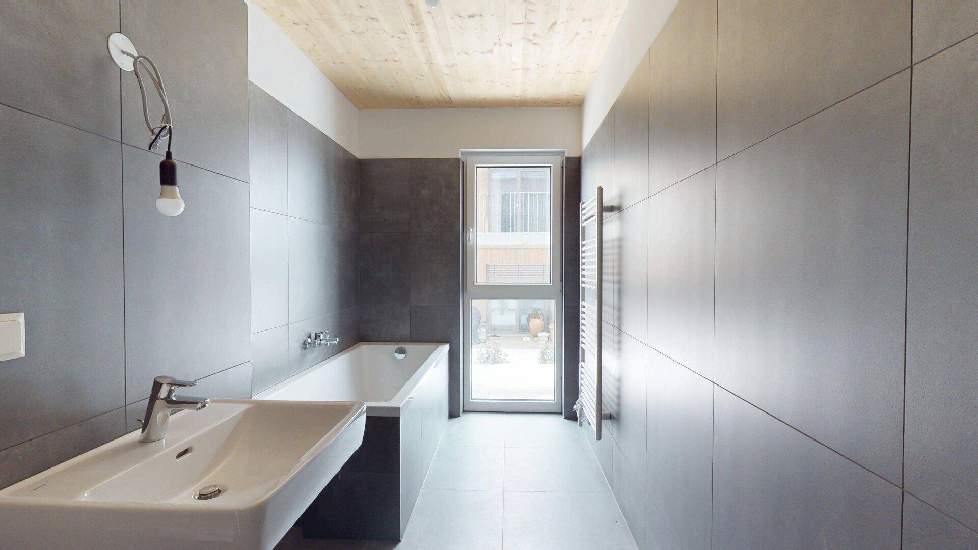 Modernes Badezimmer mit grauen Fliesen, Badewanne, Einzelwaschbecken und beheiztem Handtuchhalter.
