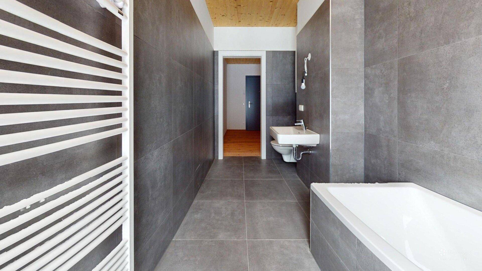 Modernes Badezimmer mit grauen Fliesen, Badewanne, Wand-WC und beheiztem Handtuchhalter.