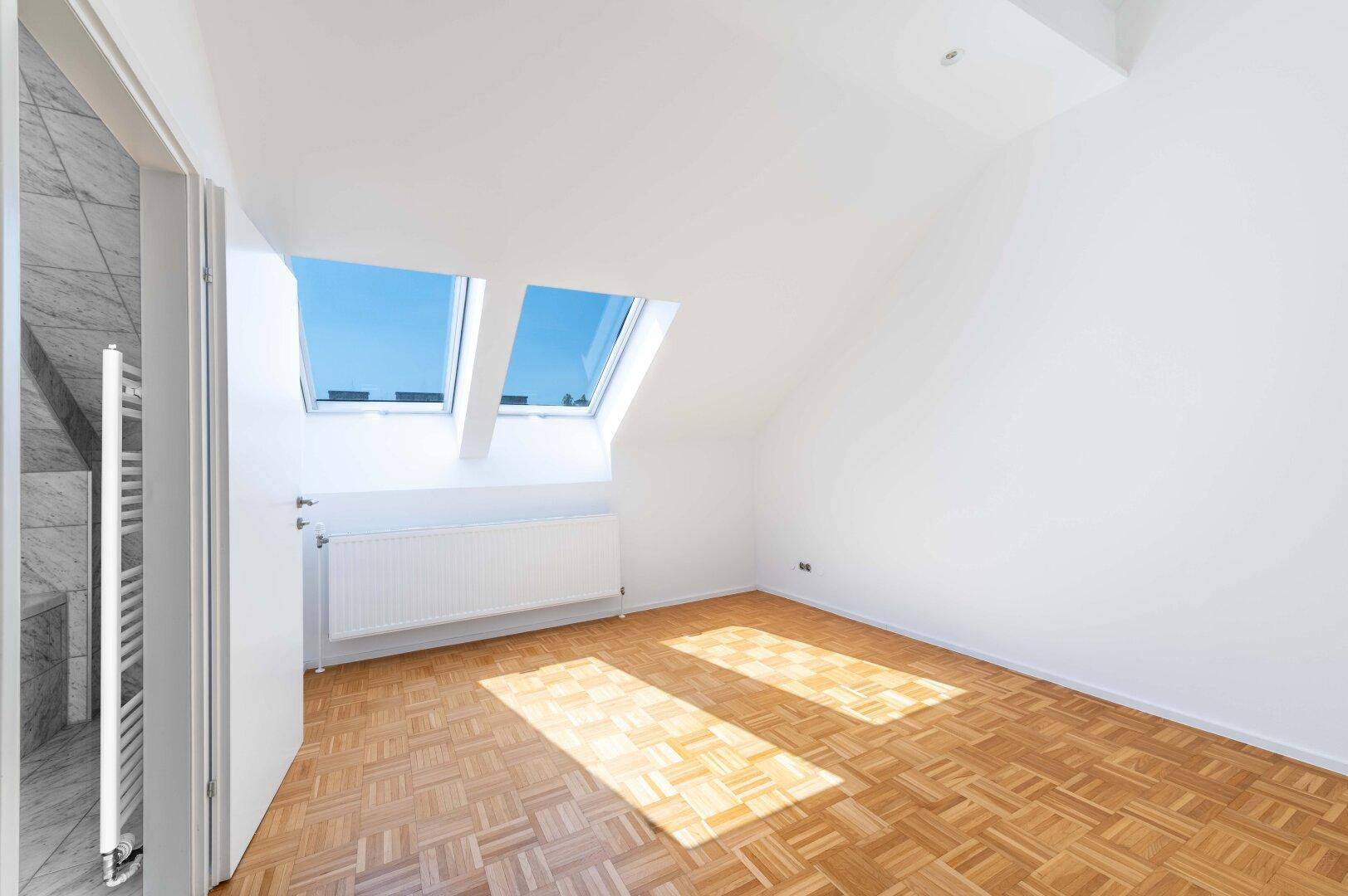 Helles Zimmer mit Parkettboden, Dachfenstern und schrägen Wänden im Dachgeschoss.