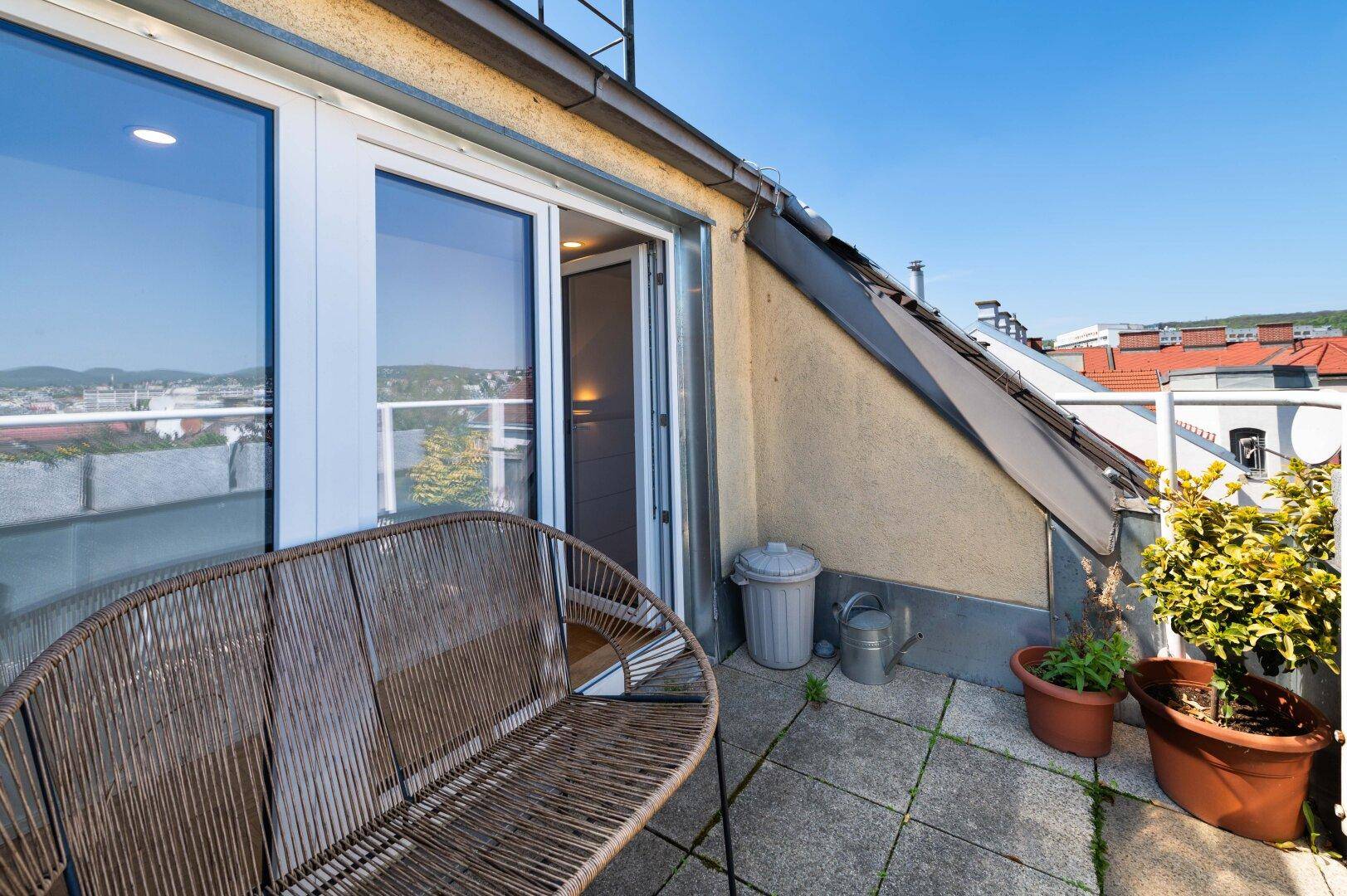 Sonnige Terrasse mit Rattanbank, Pflanzen und Blick über die Dächer der Stadt.