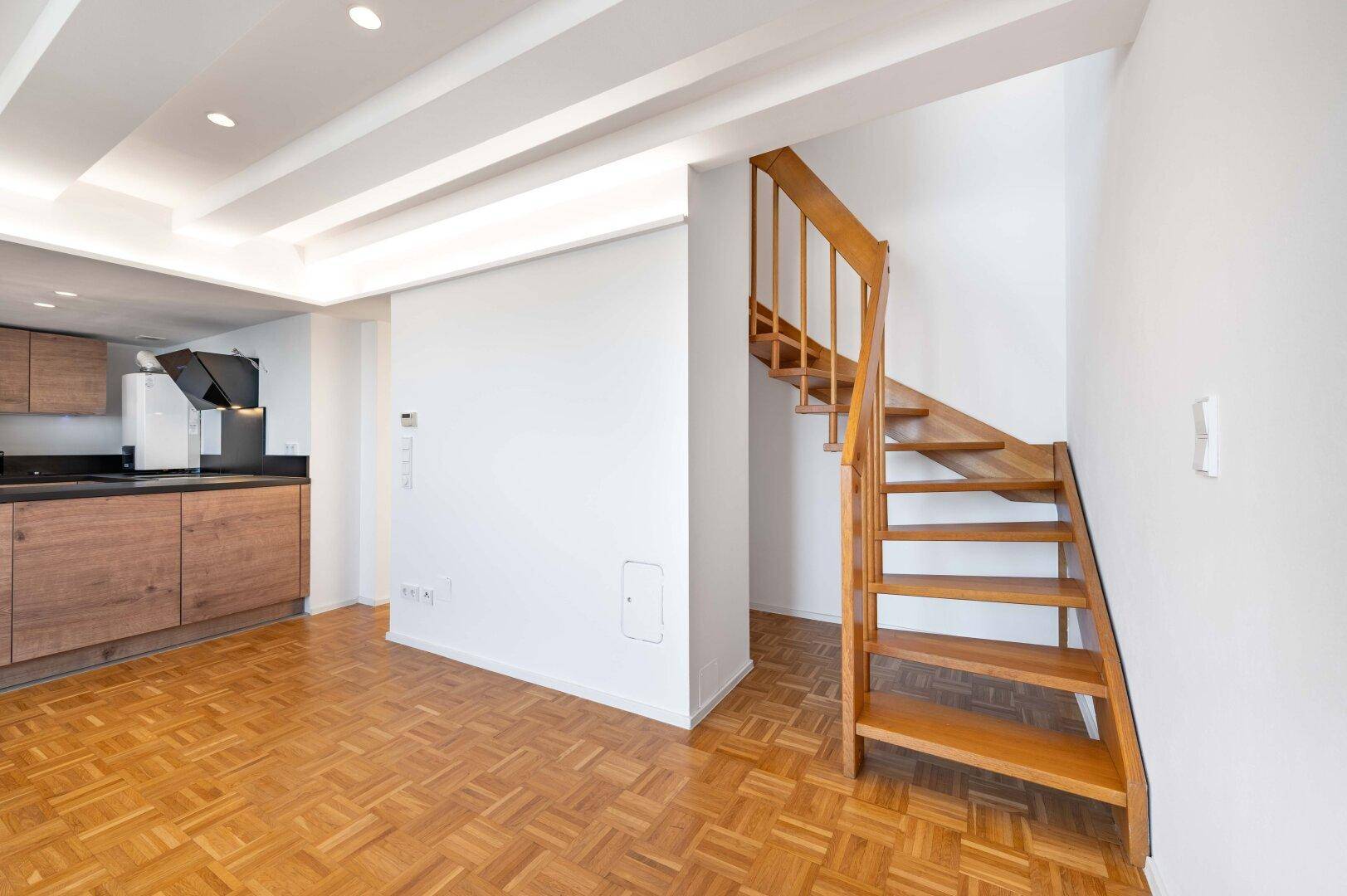 Offener Wohnbereich mit Holztreppe, Parkettboden und integrierter Küche im Hintergrund.