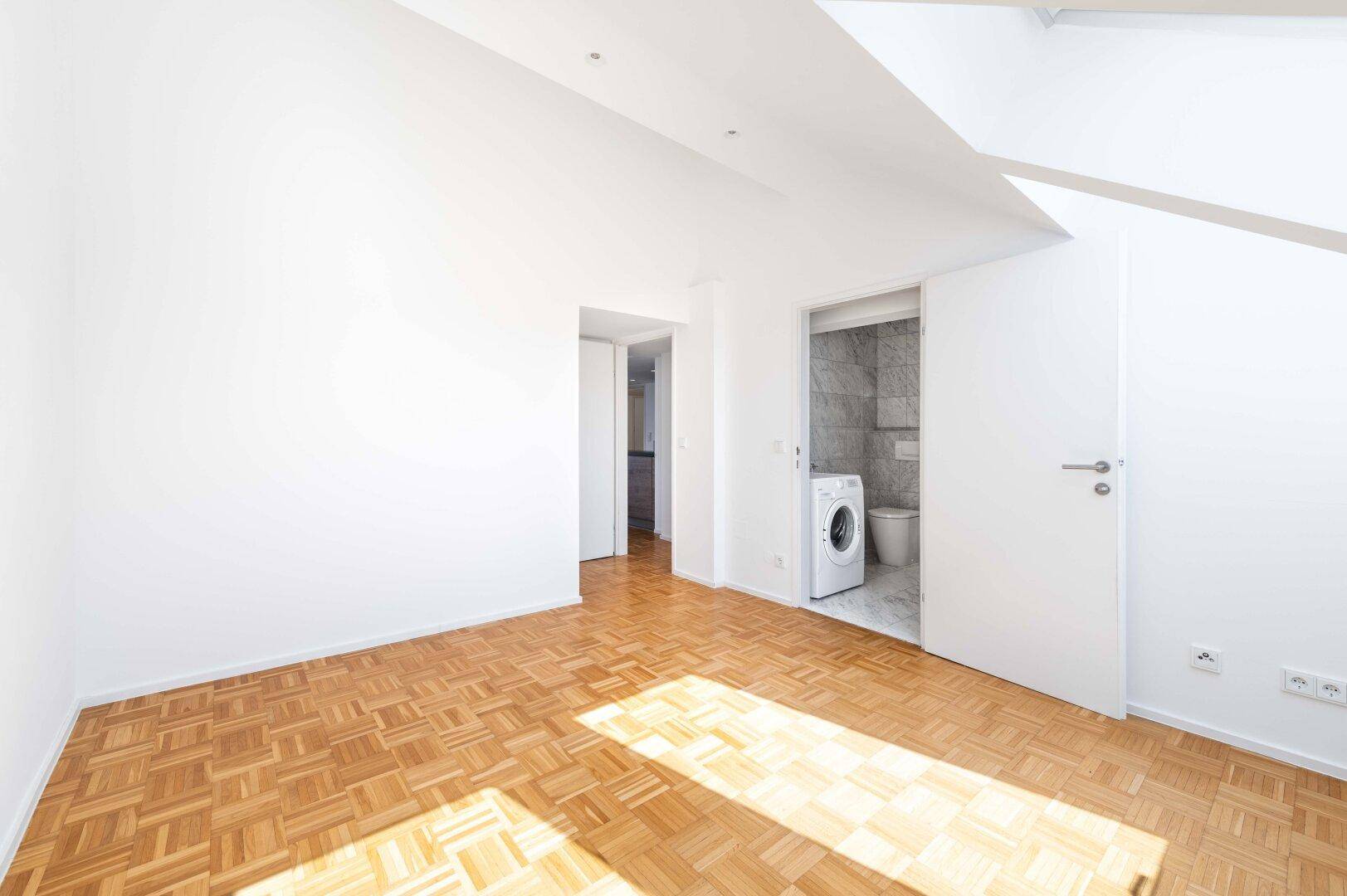 Geräumiges Zimmer mit Parkettboden und Zugang zu einem Badezimmer mit Waschmaschine.