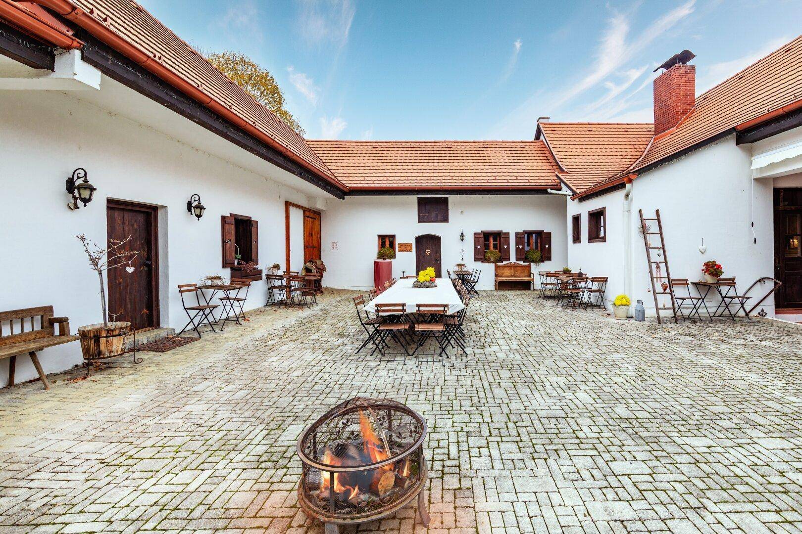 Gemütlicher Innenhof mit Feuerstelle, langen Tischen und traditioneller Architektur unter blauem Himmel.