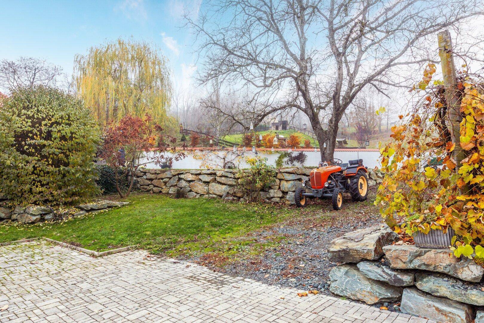 Gartenbereich mit Steineinfassung, Rasenfläche und einem alten Traktor unter Bäumen.
