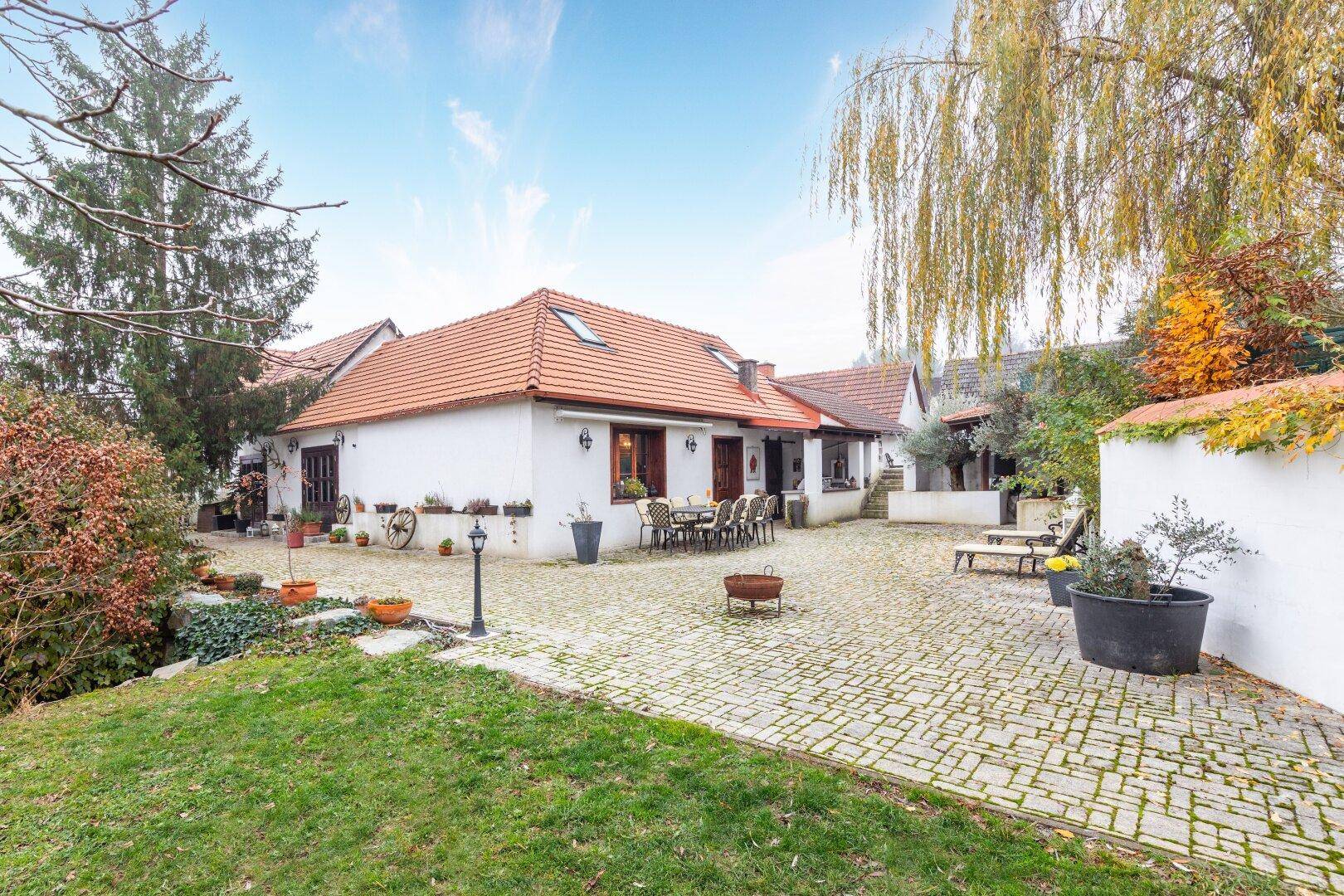 Weitläufiger gepflasterter Innenhof mit Sitzgelegenheiten, umgeben von Grünflächen und einem Weidenbaum.