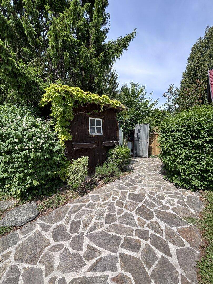 Rustikaler Holzschuppen im Garten, umgeben von üppigem Grün und einem Steinweg.