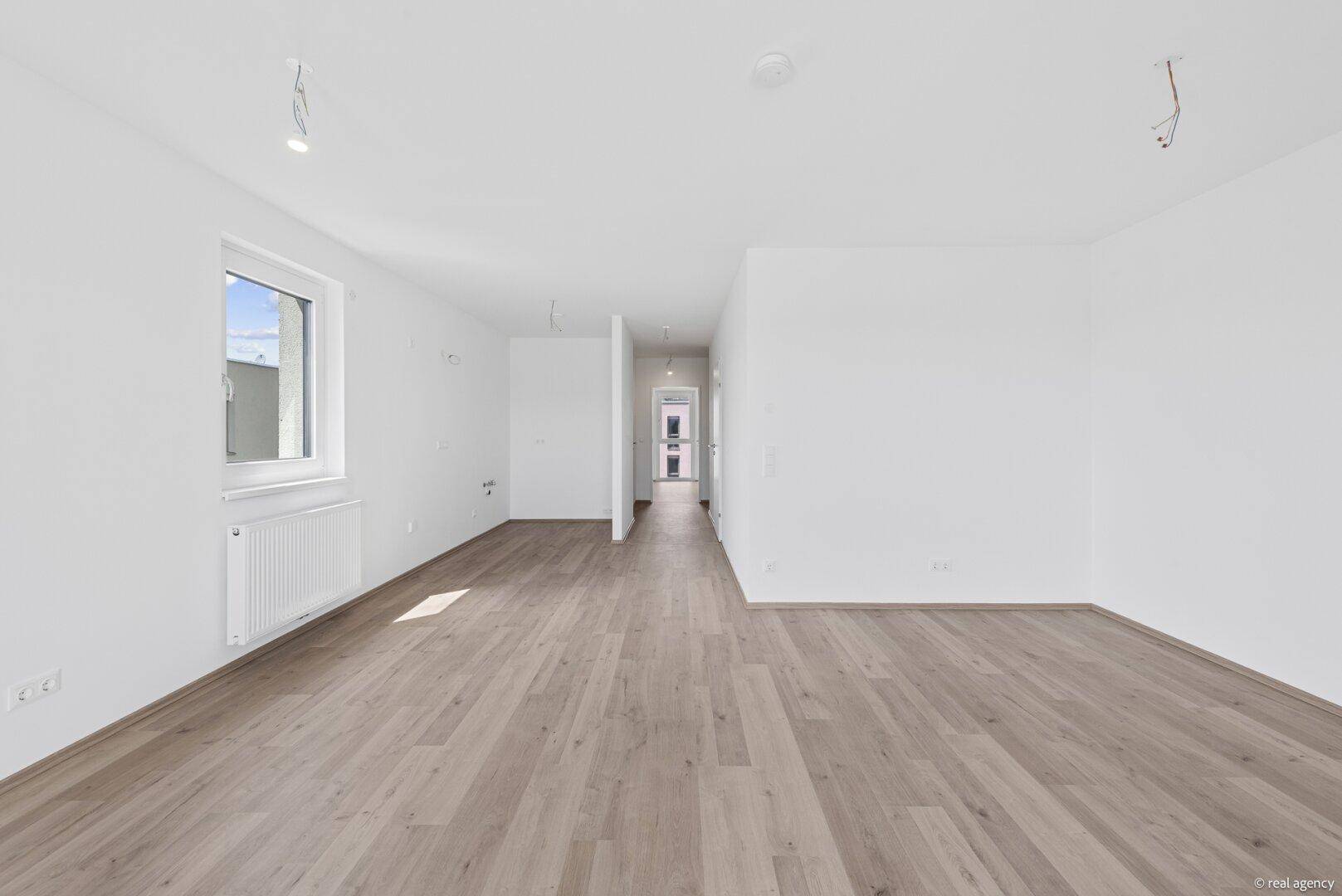 Heller, leerer Raum mit Holzboden und einem Fenster, das Tageslicht hereinlässt.