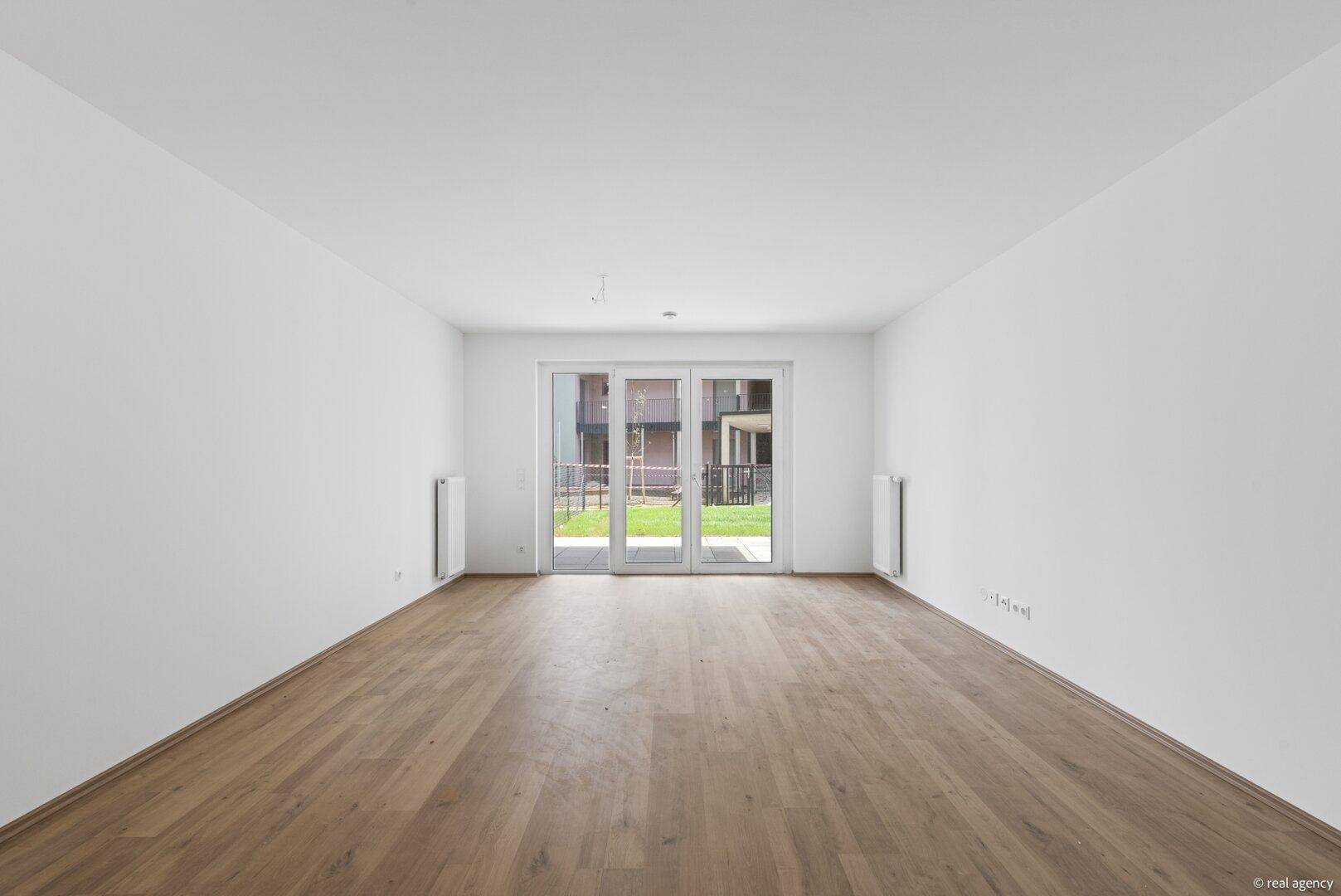 Geräumiger, leerer Wohnbereich mit Holzboden und großen Fenstertüren, die viel Tageslicht hereinlassen.