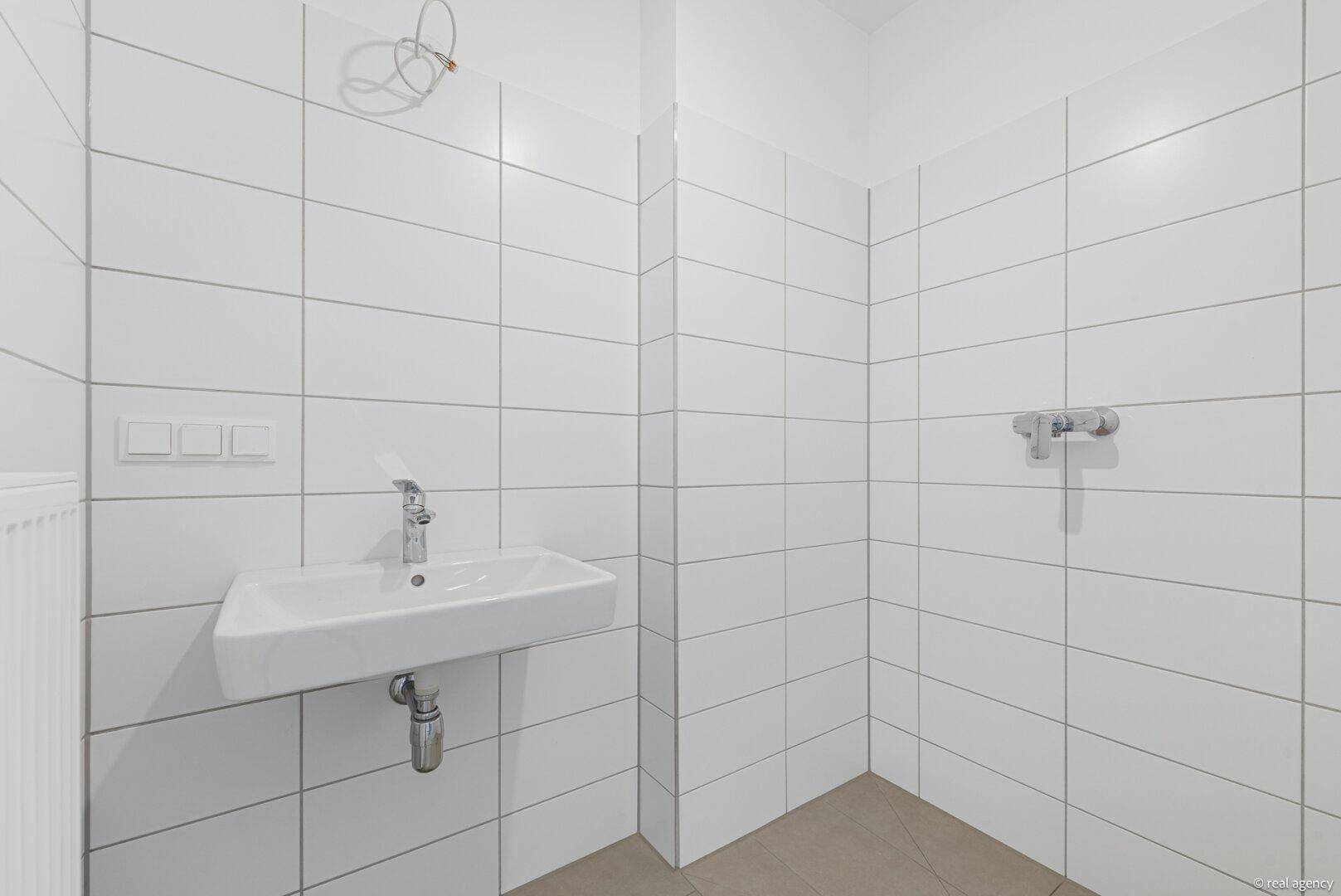 Unmöbliertes Badezimmer mit weißen Wandfliesen und vorbereiteten Anschlüssen für ein Waschbecken und eine Dusche.