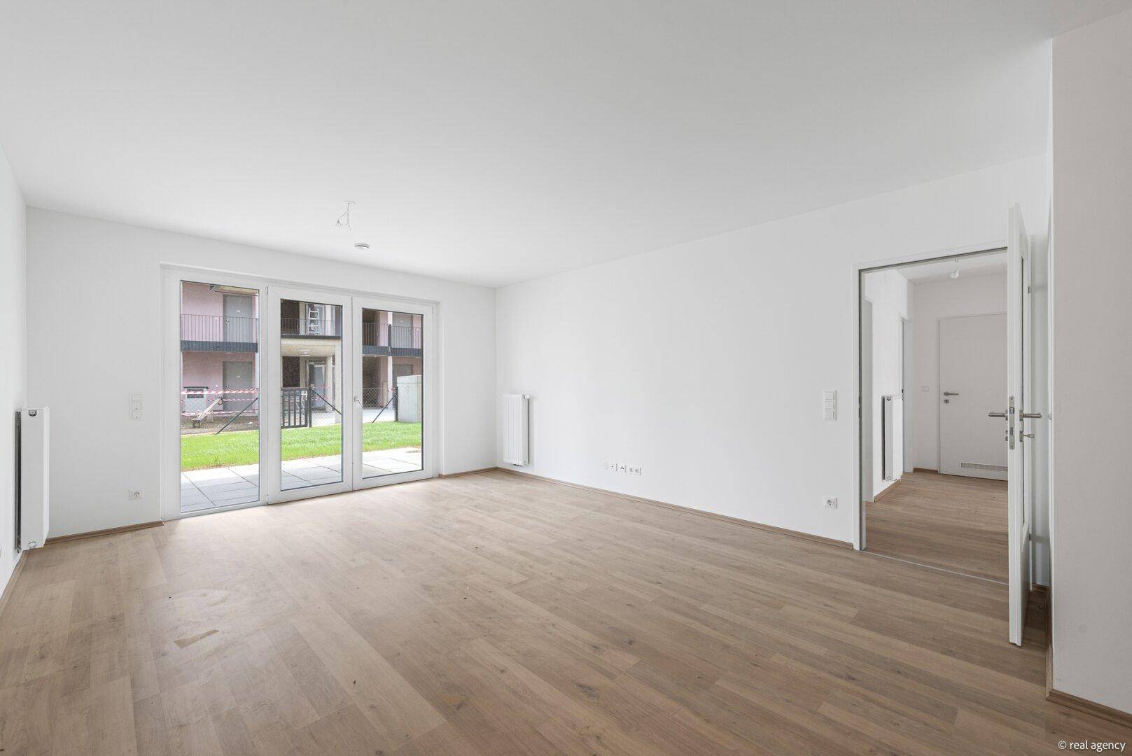 Geräumiges, unmöbliertes Wohnzimmer mit hellem Holzboden und direktem Zugang zum Gartenbereich.