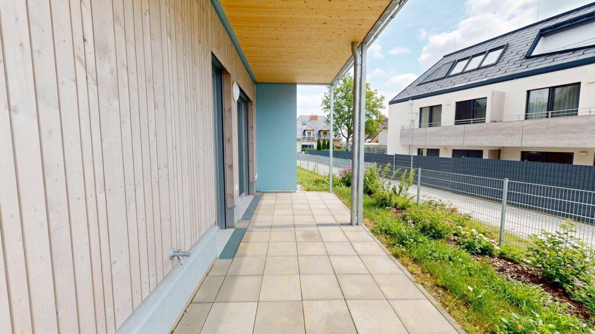 Helle Terrasse mit Holzfassade, Fliesenboden und Blick auf moderne Nachbargebäude mit Dachfenstern.