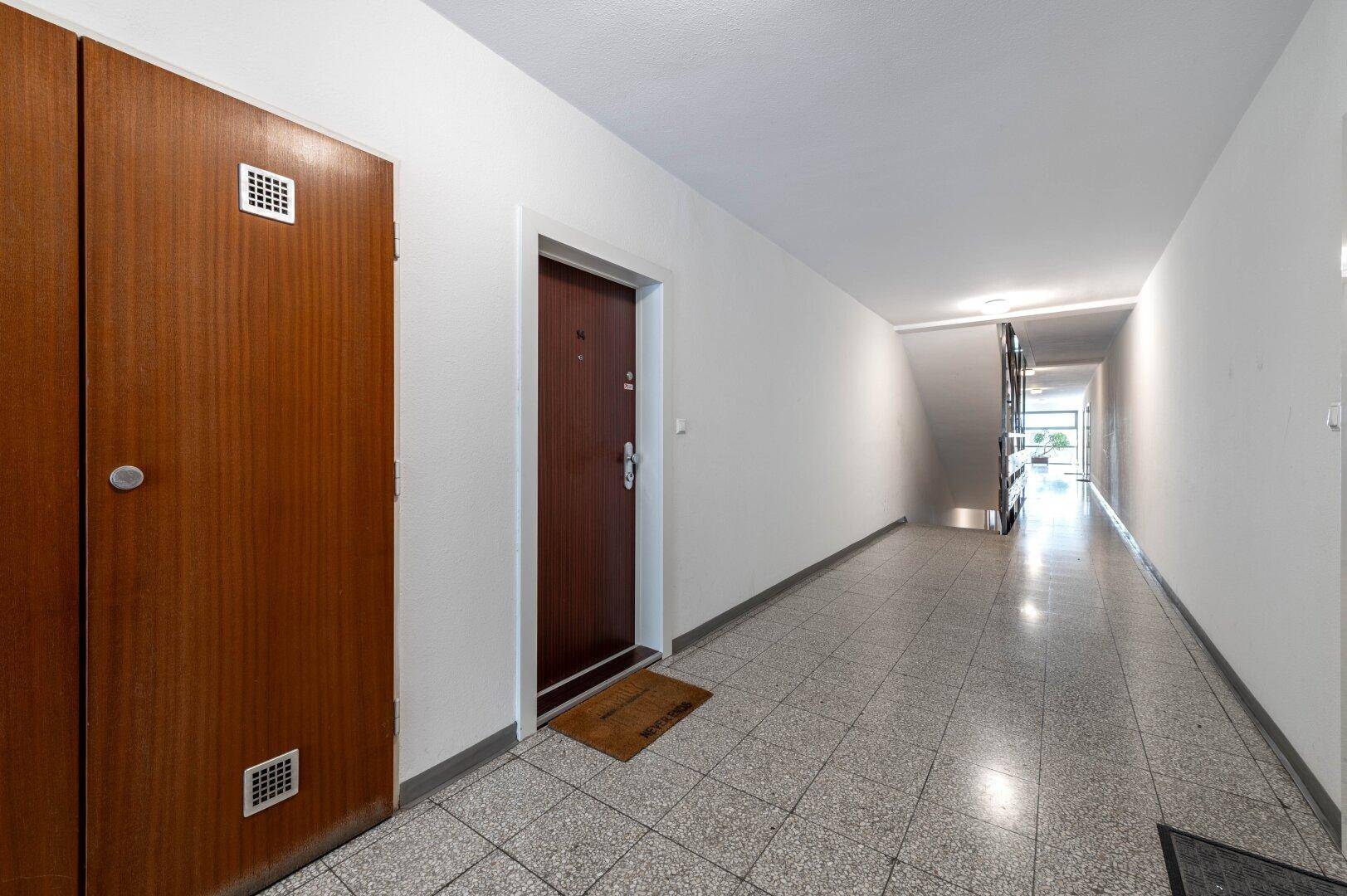 Langer Flur im Mehrfamilienhaus mit Fliesenboden und Zugang zu den Wohnungen.
