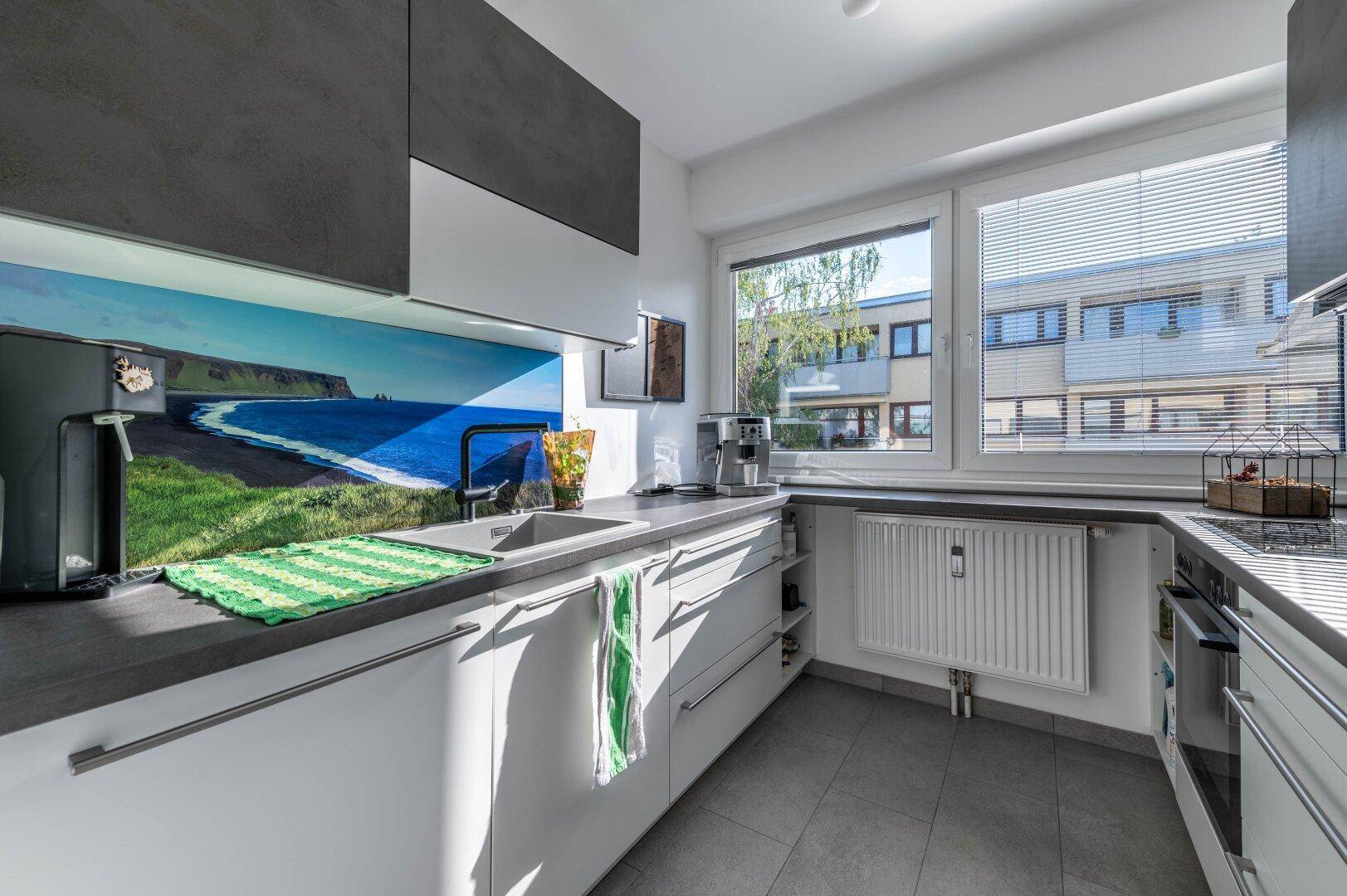 Moderne Küche mit grauen Arbeitsplatten, weißen Schränken und Fensterblick.