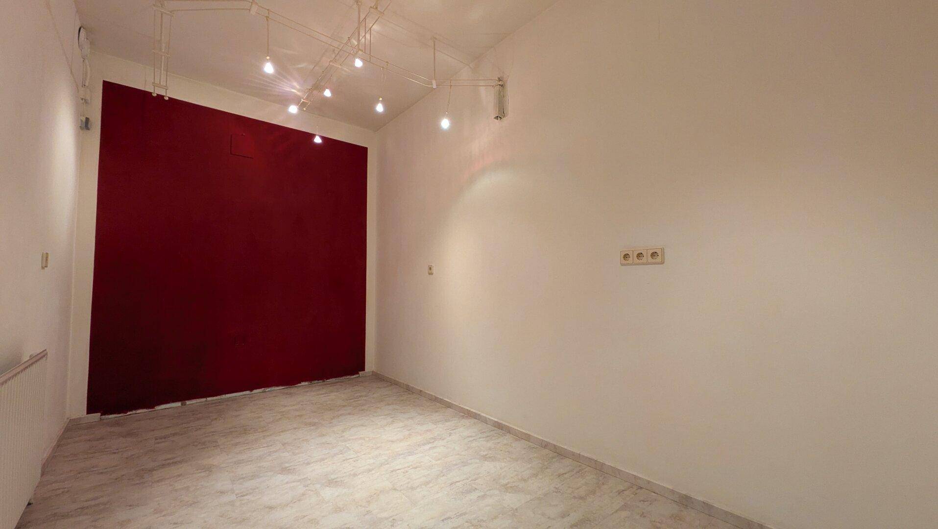 Leerer Raum mit roter Akzentwand, Fliesenboden und Schienenbeleuchtung, bereit zur Nutzung.