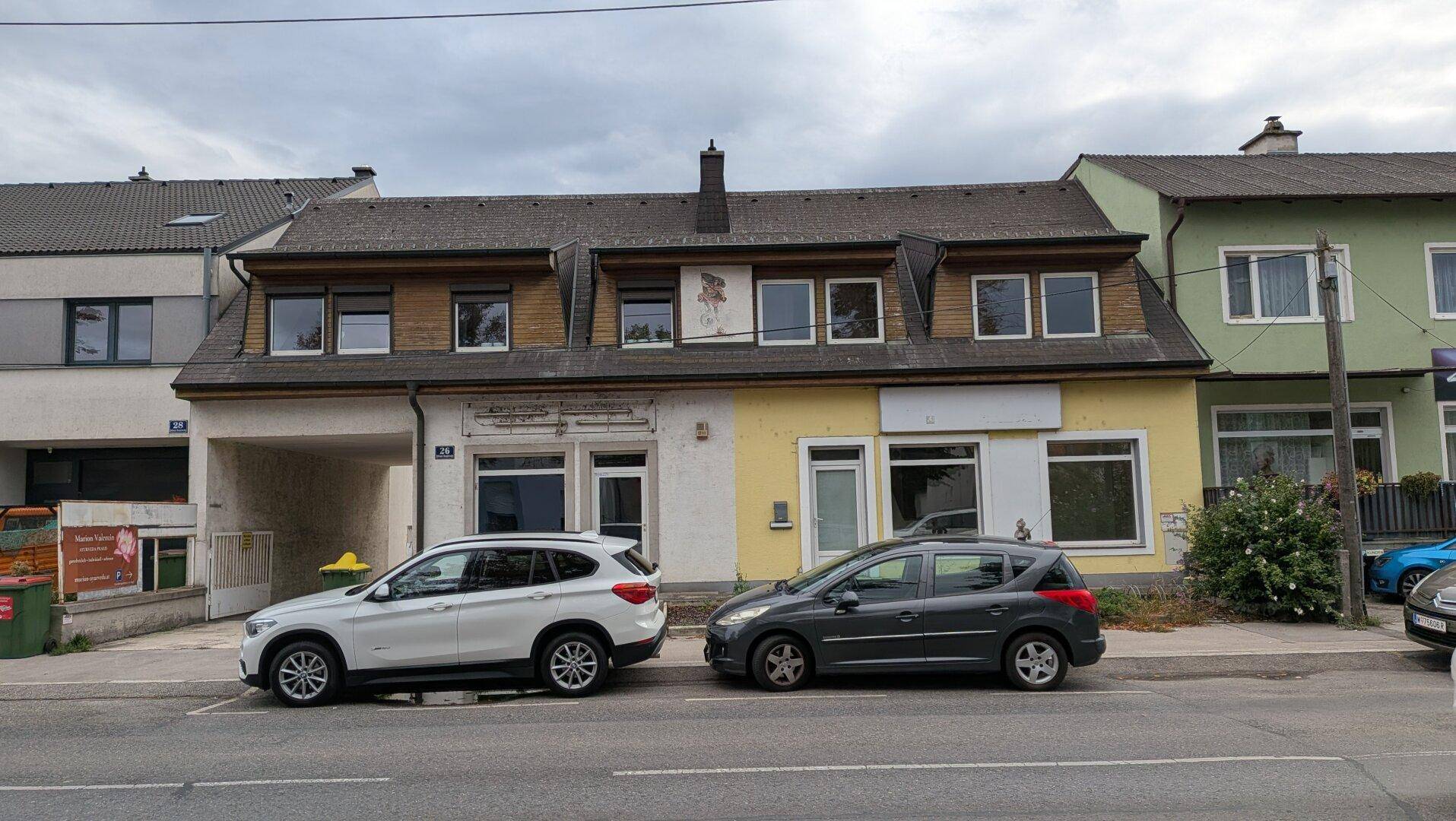 Gesamtansicht eines Gebäudes mit Geschäftsflächen im Erdgeschoss und Wohnungen, an einer Straße mit parkenden Autos.