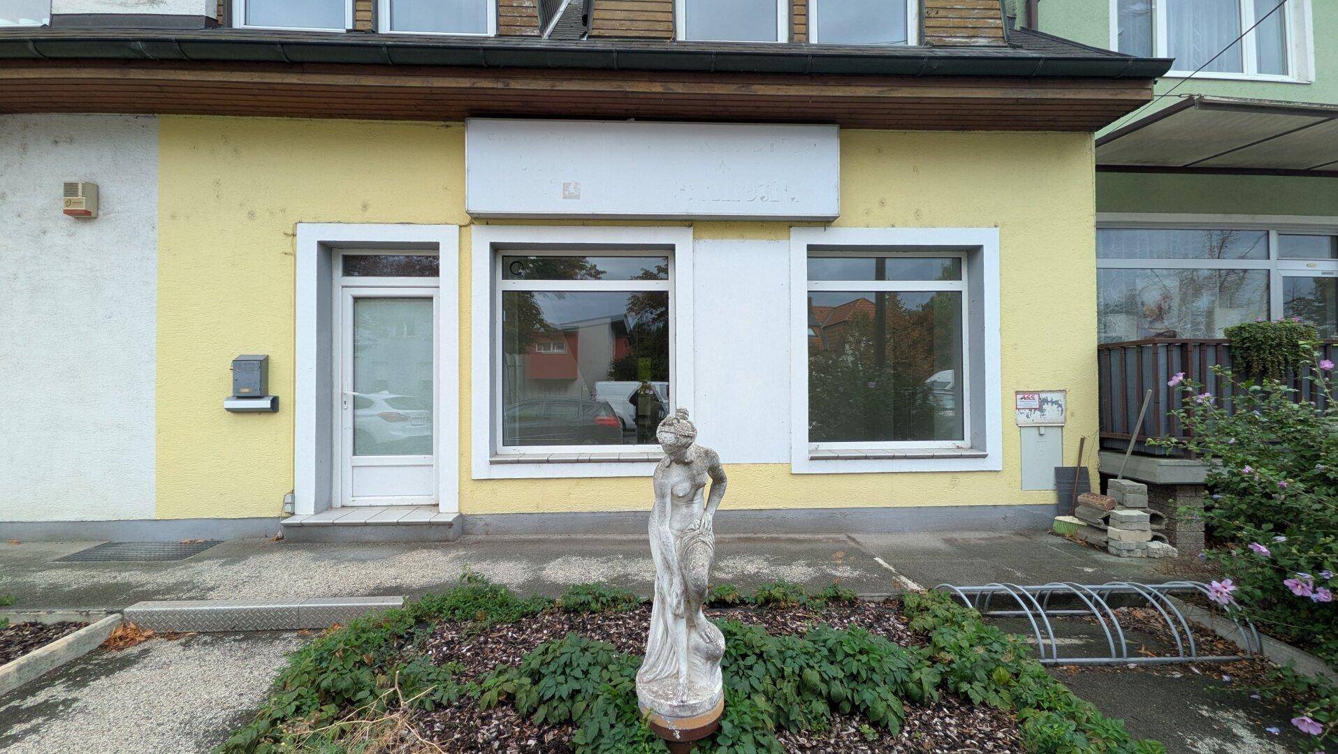 Fassade eines Geschäftsgebäudes mit leerem Schild, einfacher Bepflanzung und einer Statue.