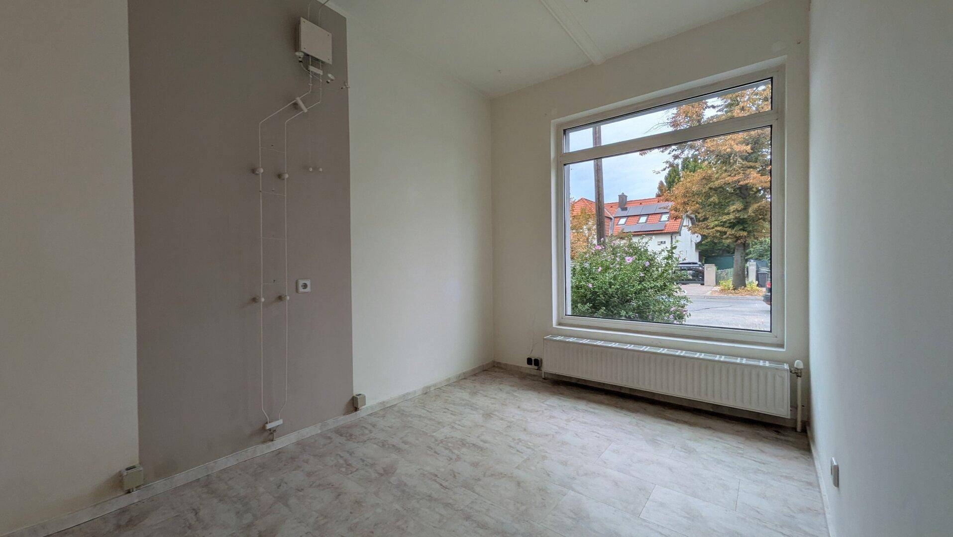 Leerer Raum mit großem Fenster, Heizkörper und einfacher Wandgestaltung, bereit zur Nutzung.