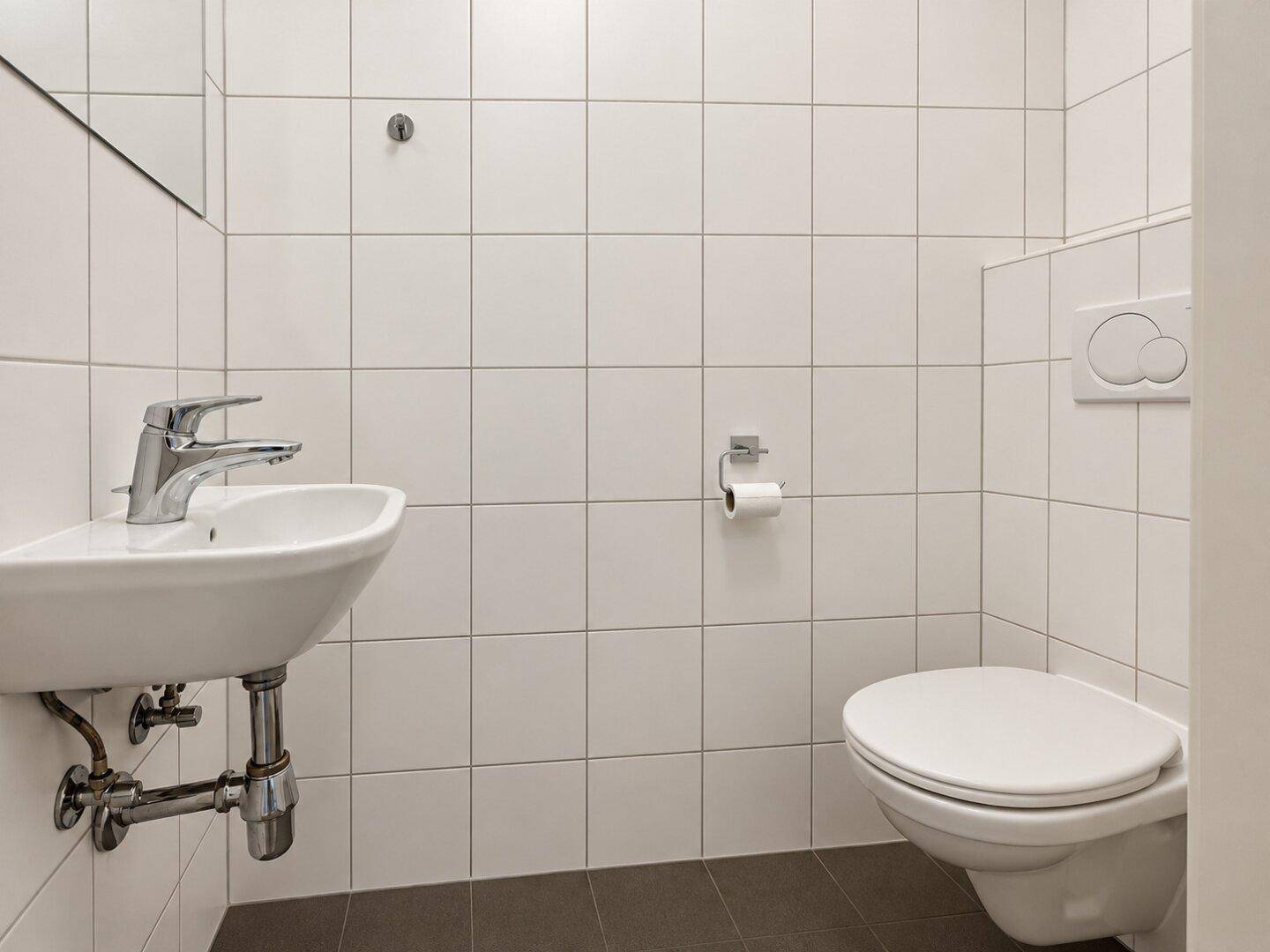 Leeres Gäste-WC mit wandhängendem WC, kleinem Waschbecken und hellen Fliesen.