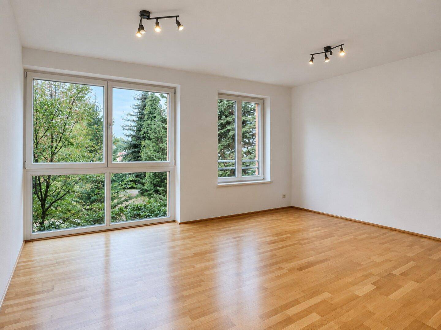 Helles, leeres Wohnzimmer mit Parkettboden und großen Fenstern mit Blick ins Grüne.