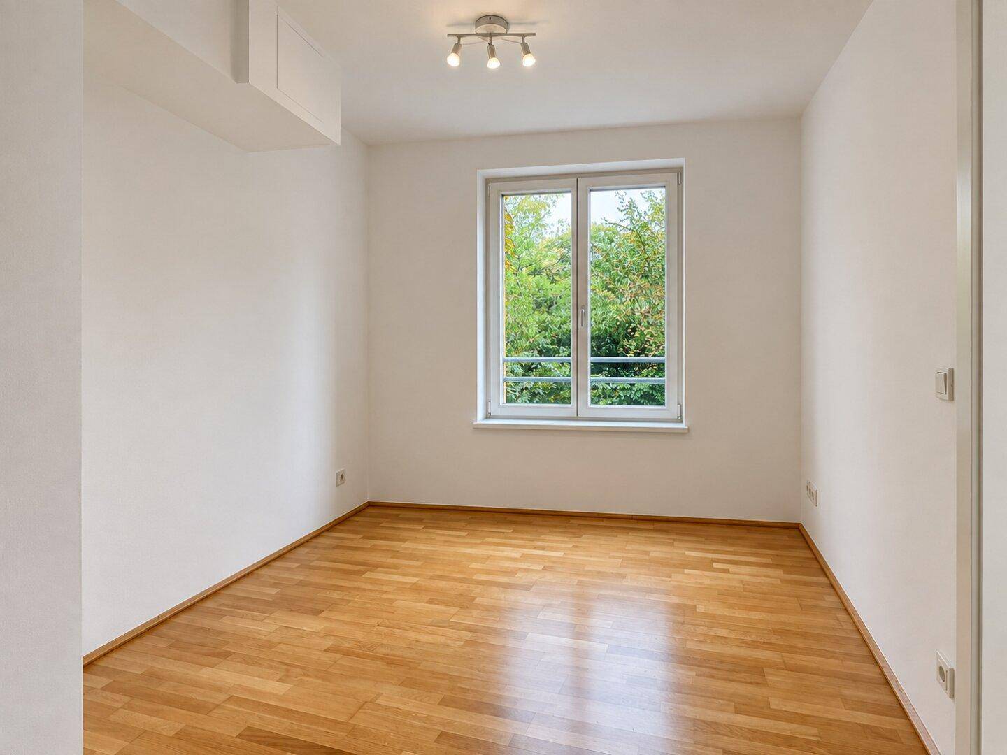 Heller, leerer Raum mit Parkettboden und einem Fenster mit Blick ins Grüne.